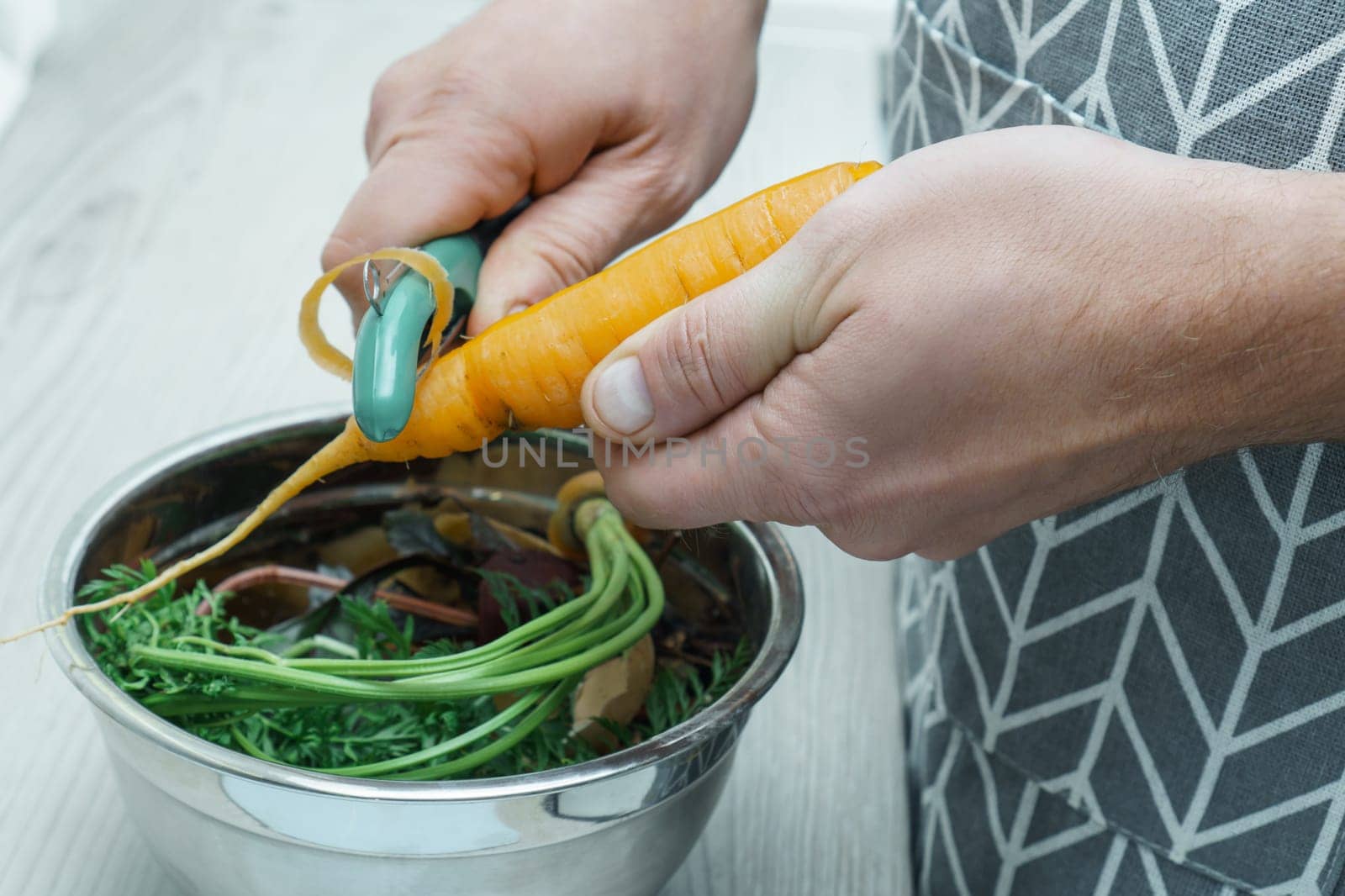 Cropped photo of man wearing grey apron peeling carrot with green peeler over metal bowl full of peelings, standing at grey wooden table in kitchen. Recycling, composting, fertilizing, zero waste.