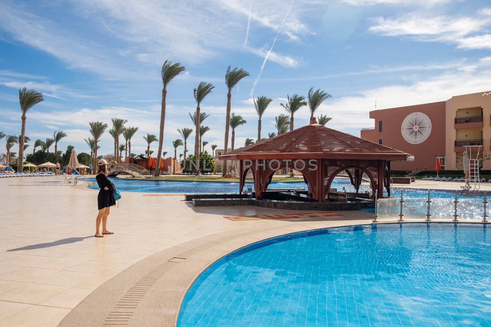 A woman stands near a swimming pool at a resort in Egypt, surrounded by palm trees and enjoying the sunny weather.