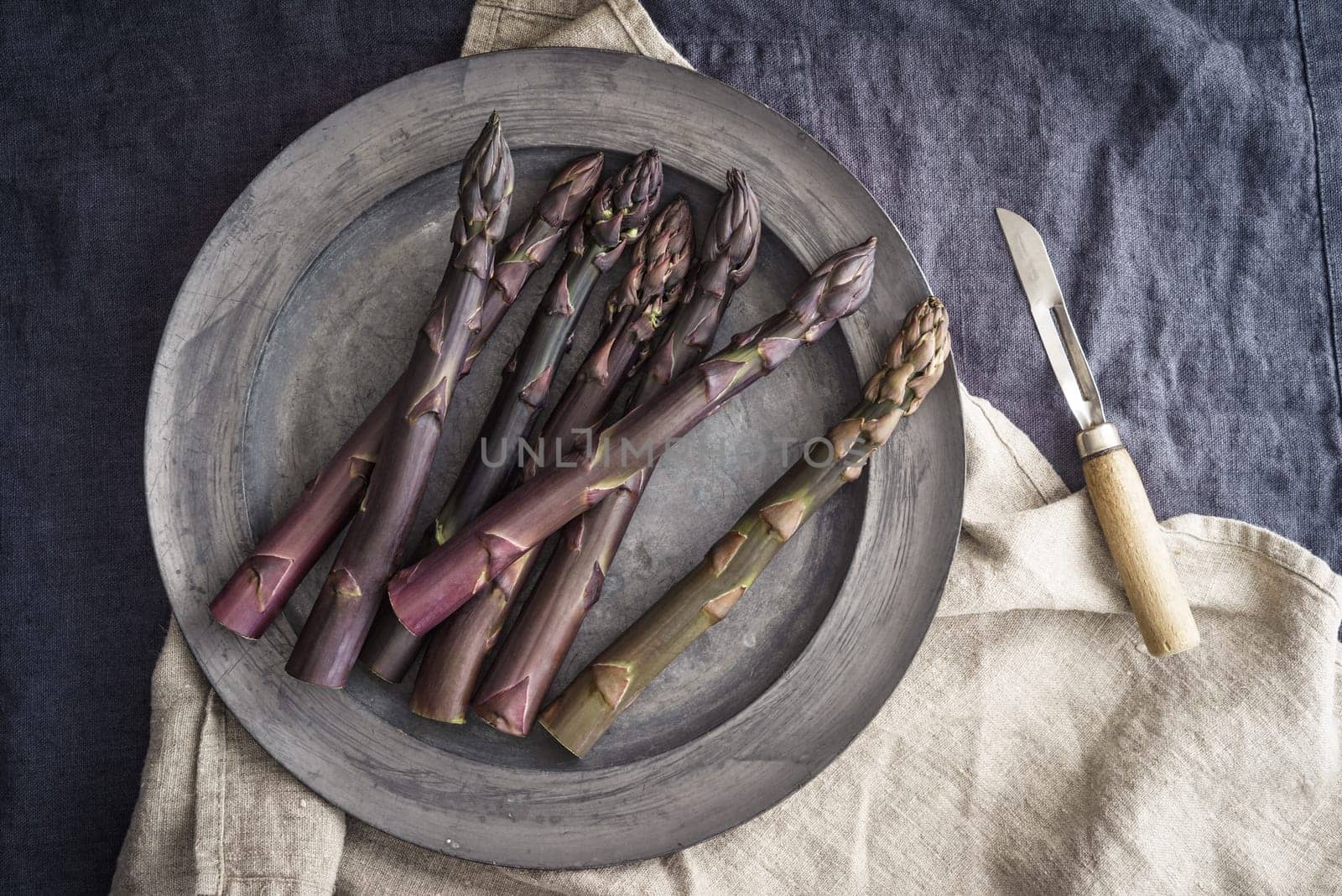 Fresh violet asparagus on old tin plate with old peeling knife on dark linen