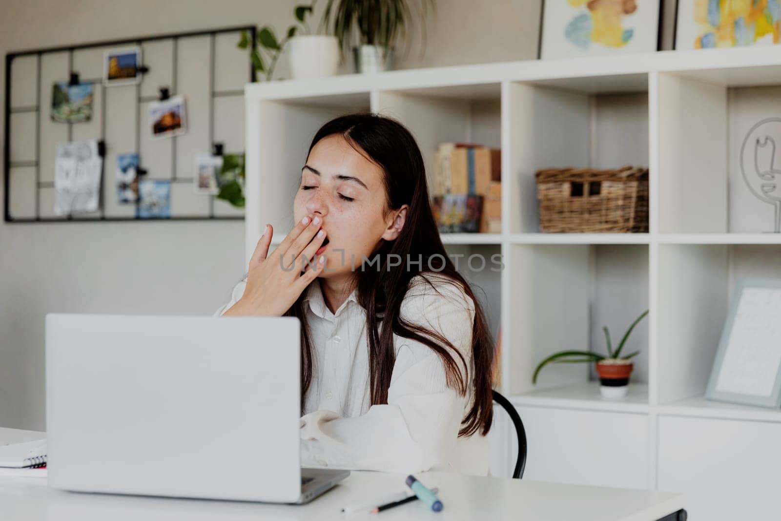 Tired brunette girl yawns tiredly with eyes closed while working with laptop at home or office. Woman is reading computer screen. Student is studying, learning. Person work hard. E-learning concept