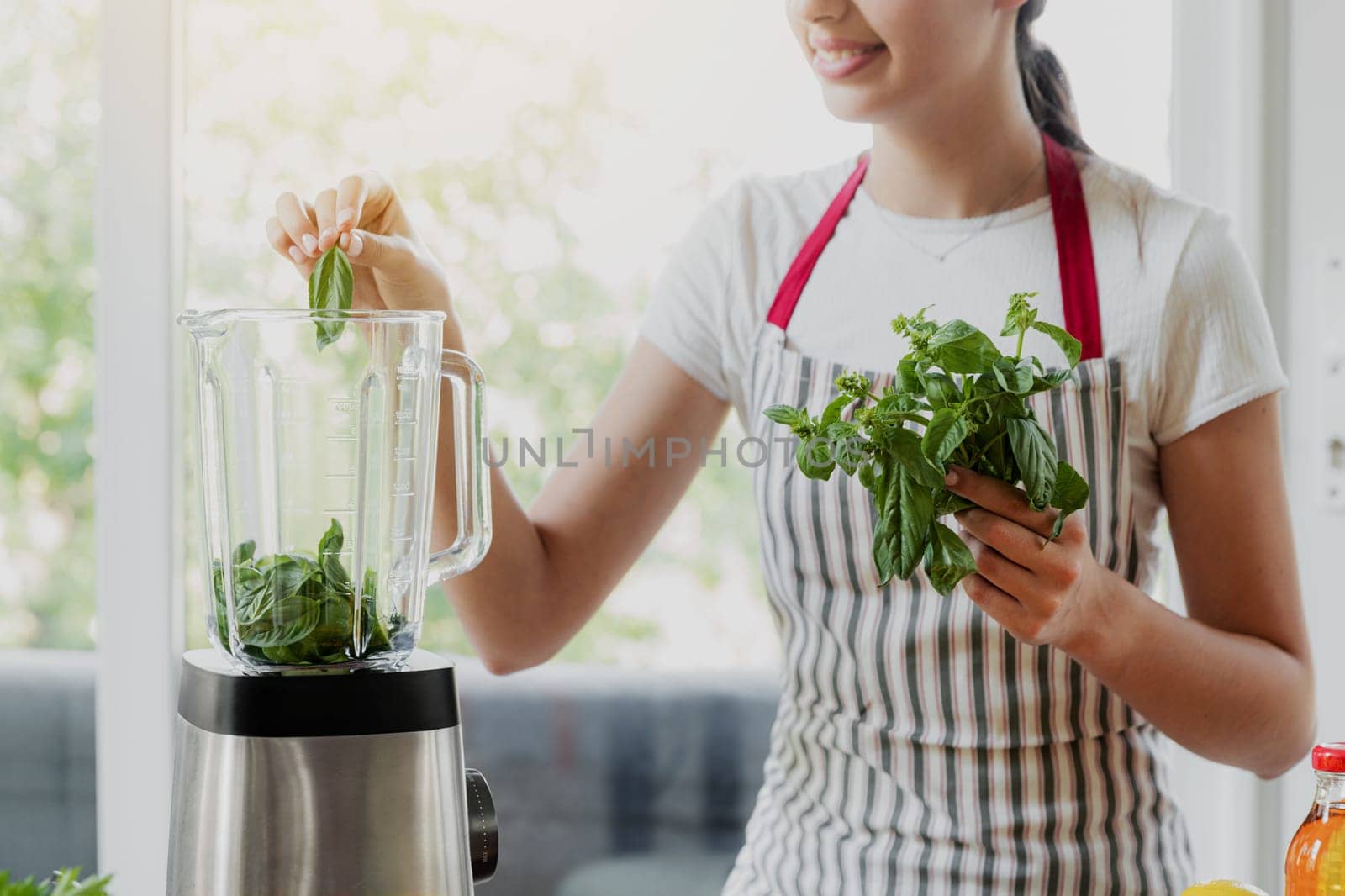 Young smiling woman making green smoothie at home kitchen with blender. Close up of beautiful girl hands put leaves in bowl for mixing. Process of healthy eating preparation, detox cocktail drink