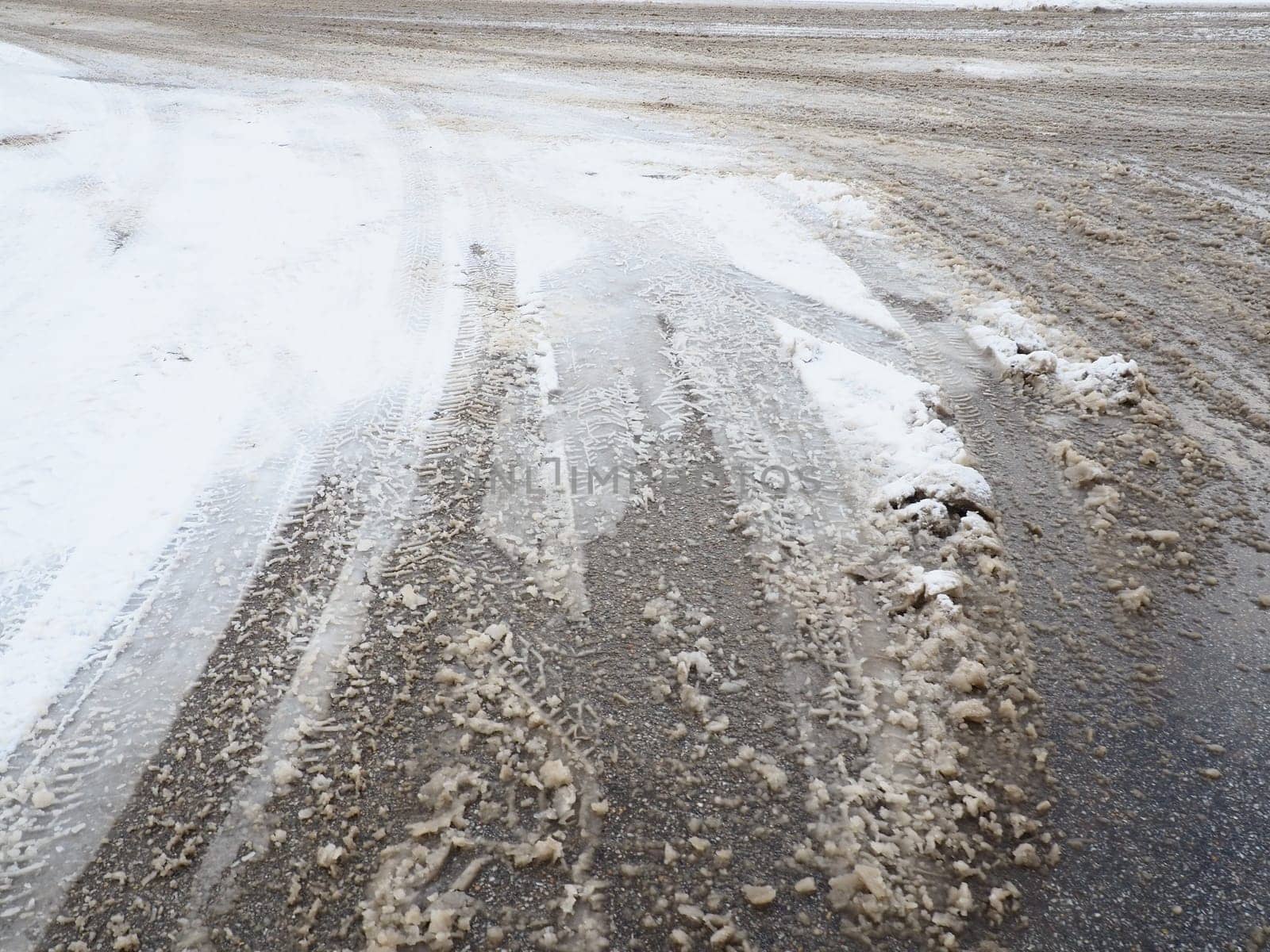 Roadside with a snowdrift. Difficult driving conditions. Wet slippery pavement with footprints. Dangerous ice. Muddy weather in winter. Car tire tracks. The work of municipal services for cleaning.