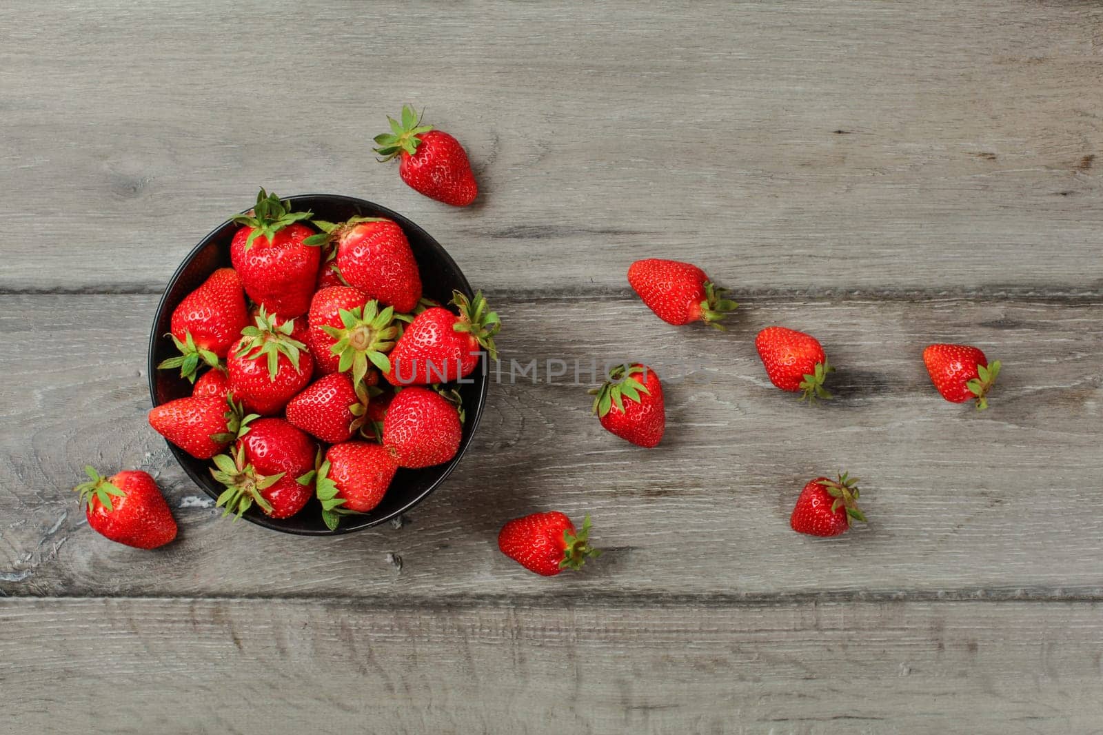 Tabletop view, small black ceramic bowl with strawberries, some of them spilled next to it on gray wood desk.