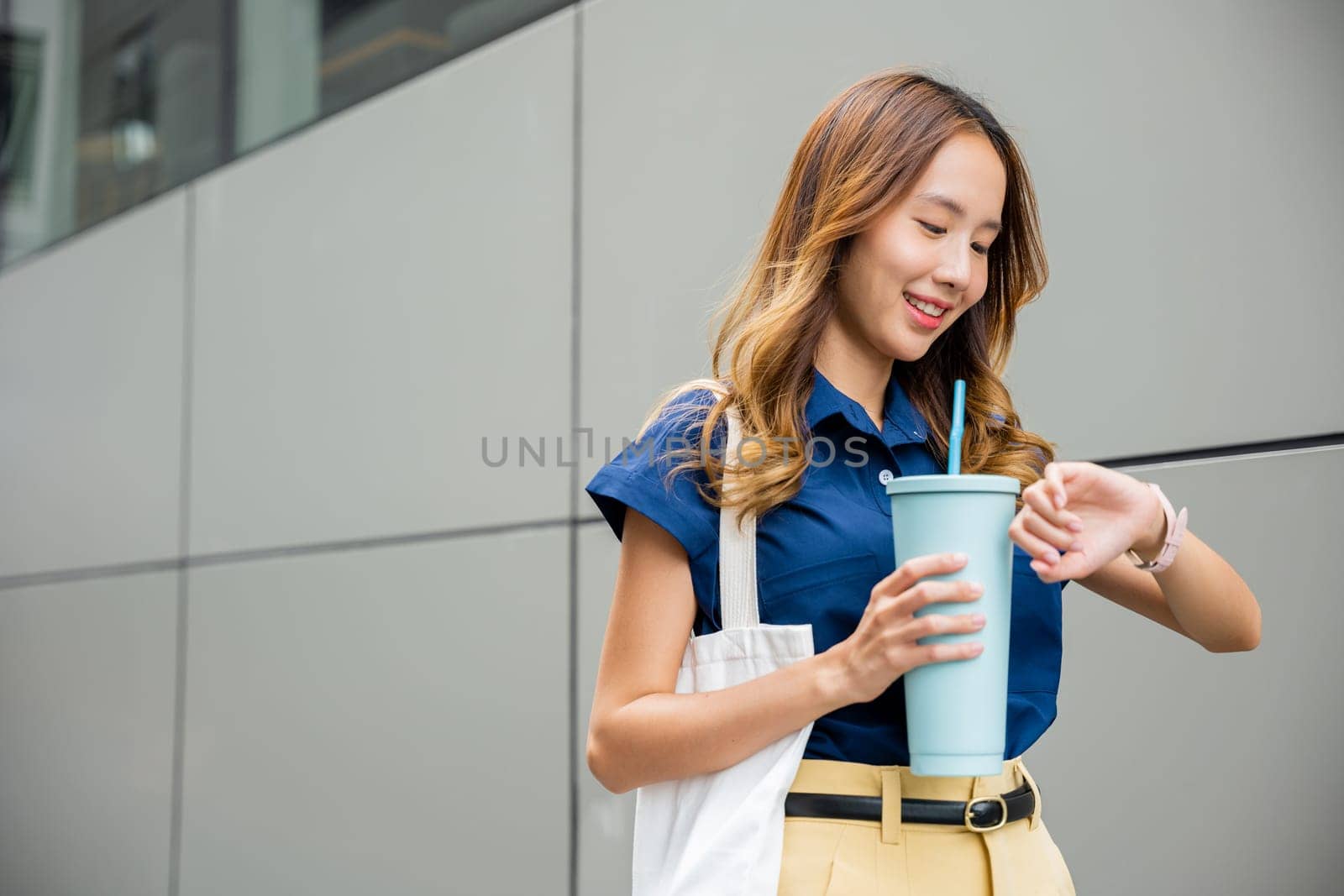 Asian beautiful woman confident smiling with cloth bag holding steel thermos tumbler mug water glass she walking outdoors on street near modern building office, Happy female looking watch time