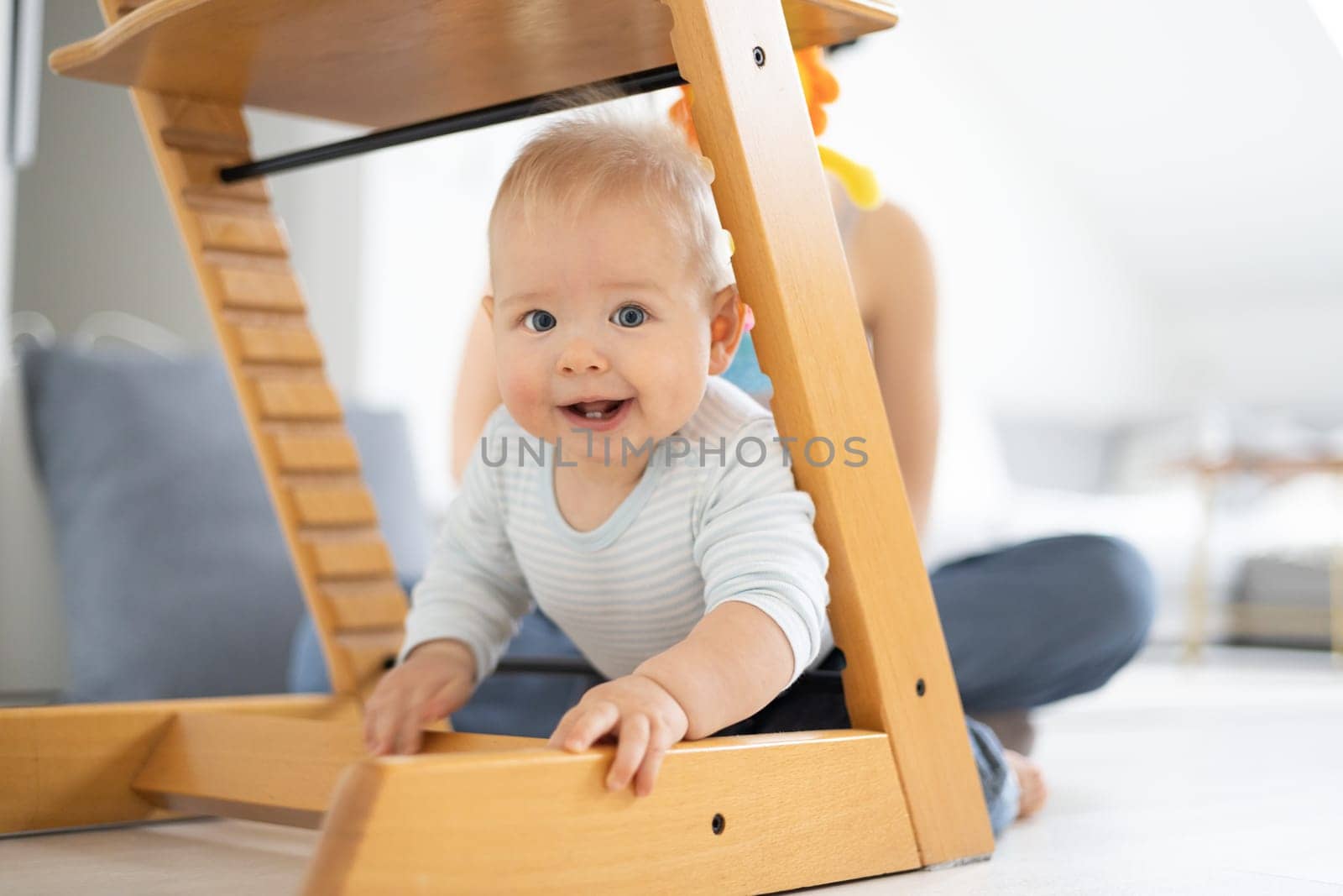 Happy infant climbing under traditional scandinavian designer wooden high chair and in modern bright home. Cute baby smiling in camera.