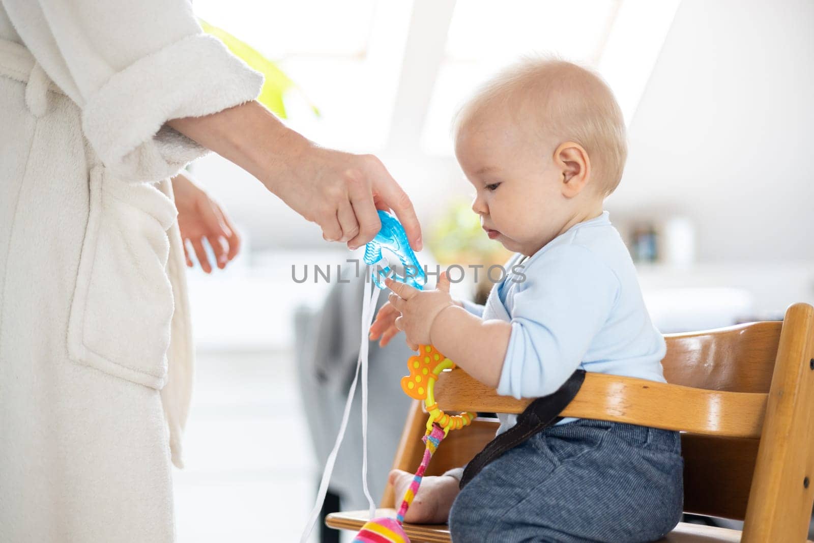 Happy infant sitting at dining table and playing with his toy in traditional scandinavian designer wooden high chair in modern bright atic home superwised by his mother.