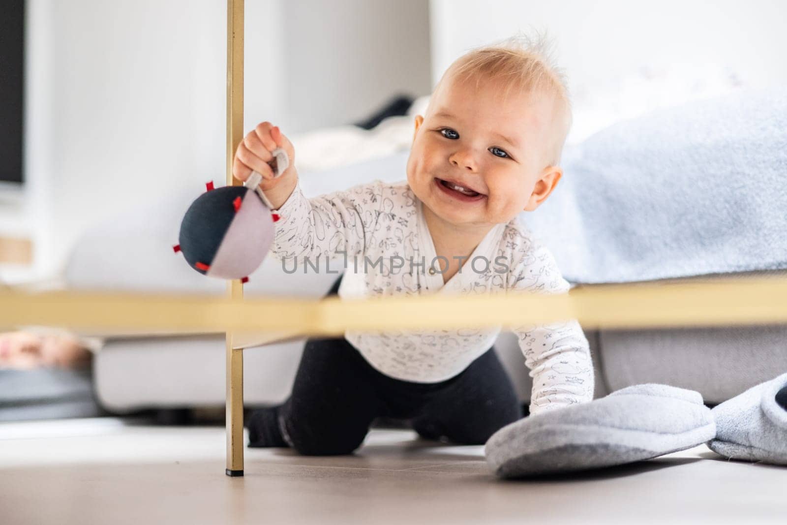 Cute infant baby boy playing with hanging ball, crawling and standing up by living room table at home. Baby activity and play center for early infant development. Baby playing at home