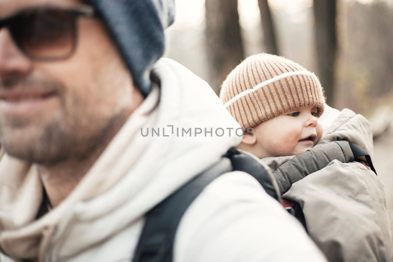 Sporty father carrying his infant son wearing winter jumpsuit and cap in backpack carrier hiking in autumn forest