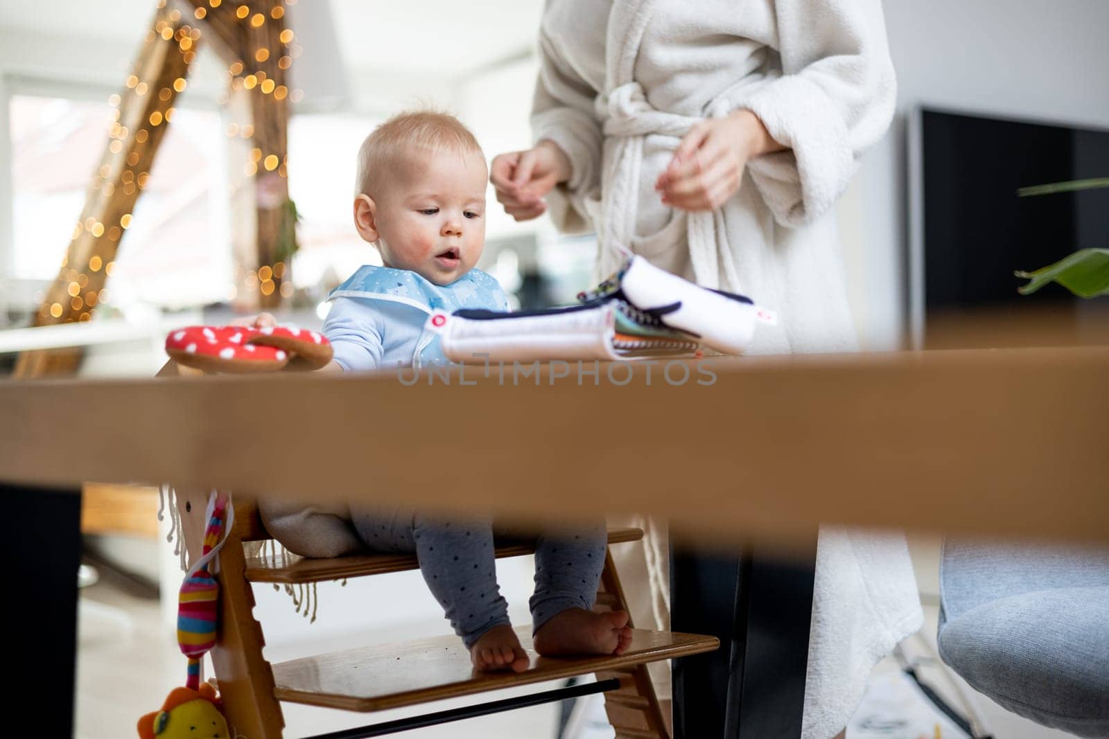 Happy infant sitting at dining table and playing with his toy in traditional scandinavian designer wooden high chair in modern bright atic home superwised by his mother.