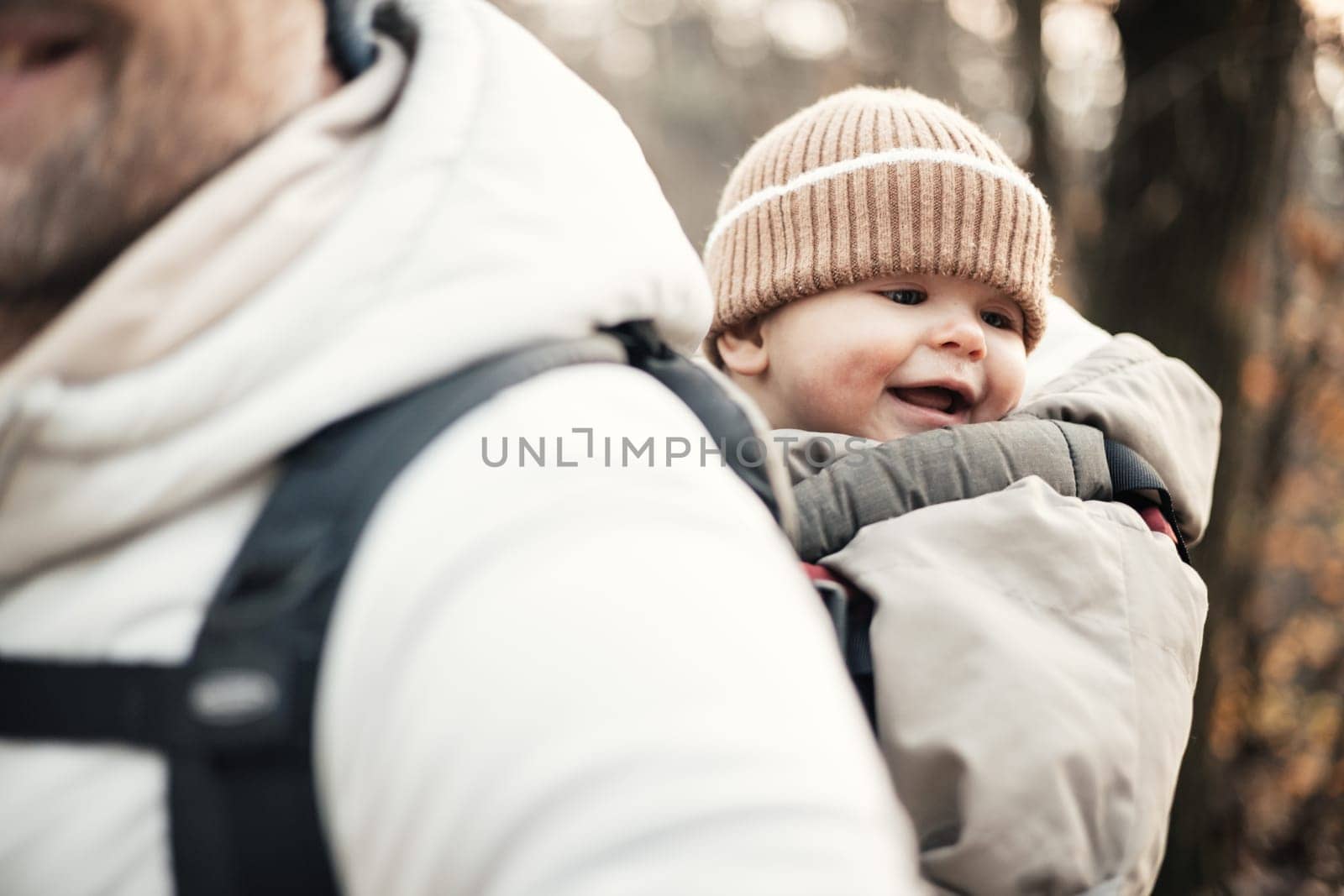 Sporty father carrying his infant son wearing winter jumpsuit and cap in backpack carrier hiking in autumn forest