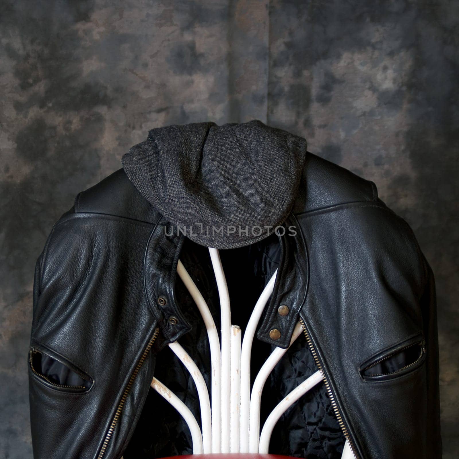 A leather biker jacket hangs on the back of a chair with a tweed cap sitting on top.