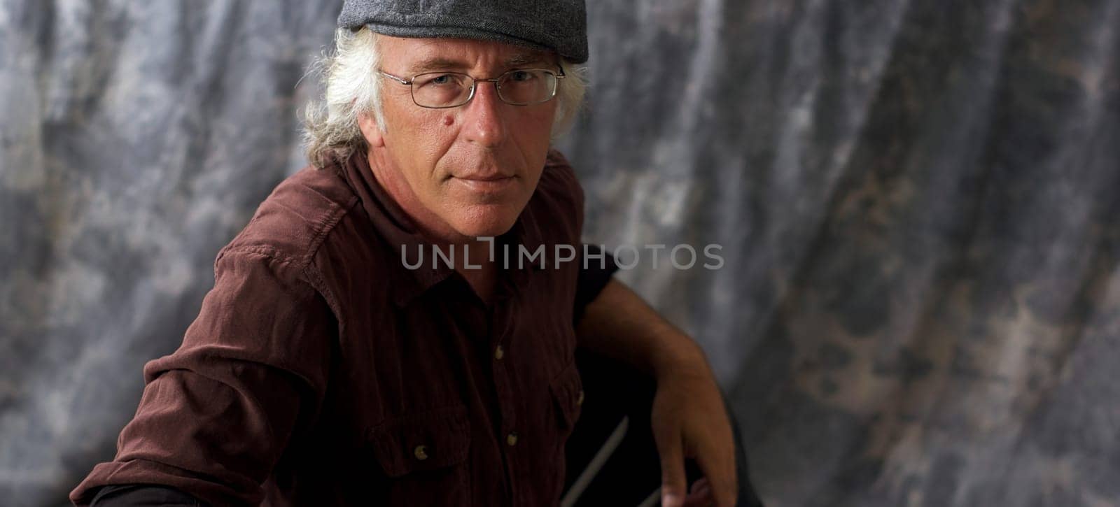 An older blue eyed man with glasses and white hair wearing a brown shirt and grey cap against mottled gray background.