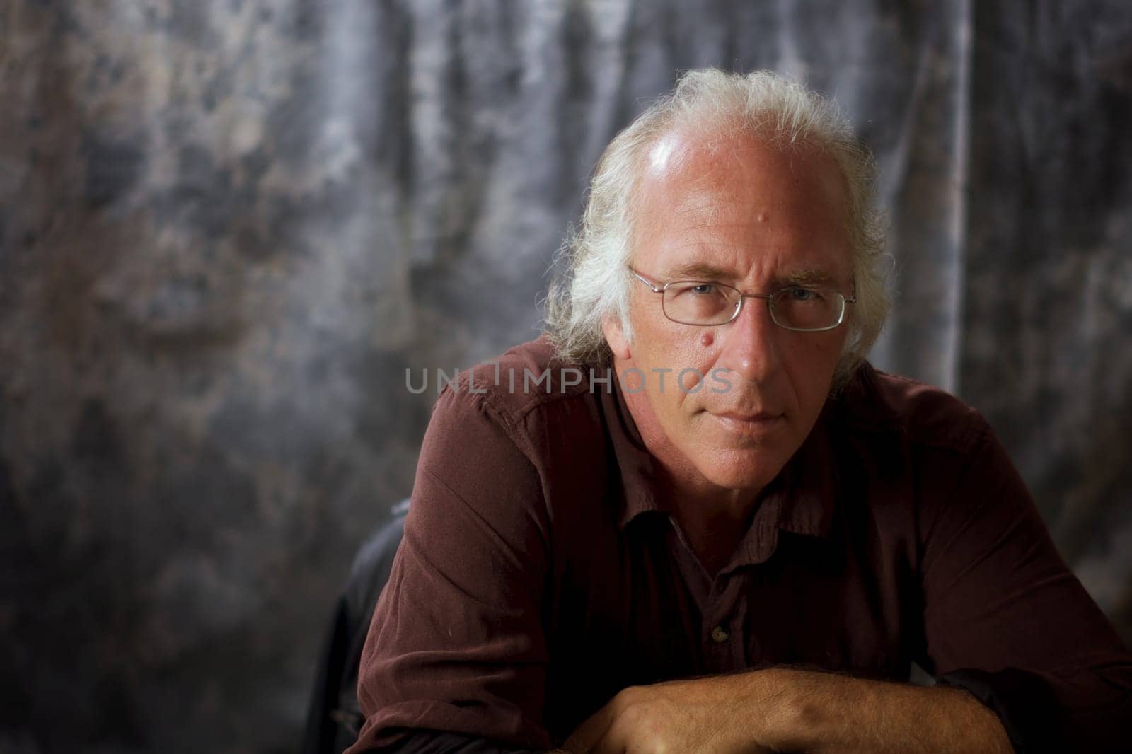 An older blue eyed man with glasses and white hair wearing a brown shirt against mottled gray background looking at viewer as if listening.