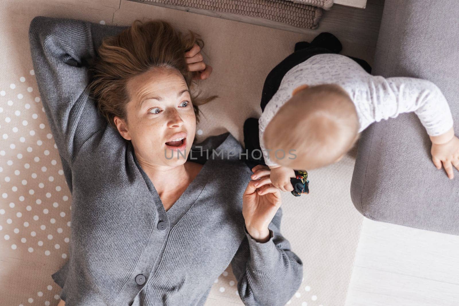 Happy family moments. Mother lying comfortably on children's mat playing with her baby boy watching and suppervising his first steps. Positive human emotions, feelings, joy