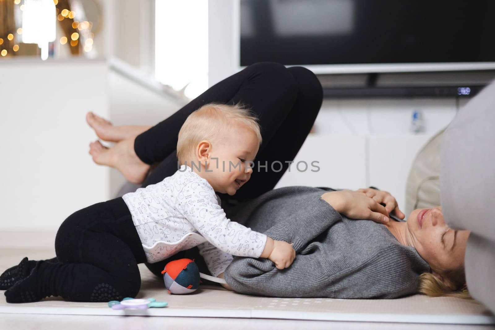 Happy family moments. Mother lying comfortably on children's mat playing with her baby boy watching and suppervising his first steps. Positive human emotions, feelings, joy