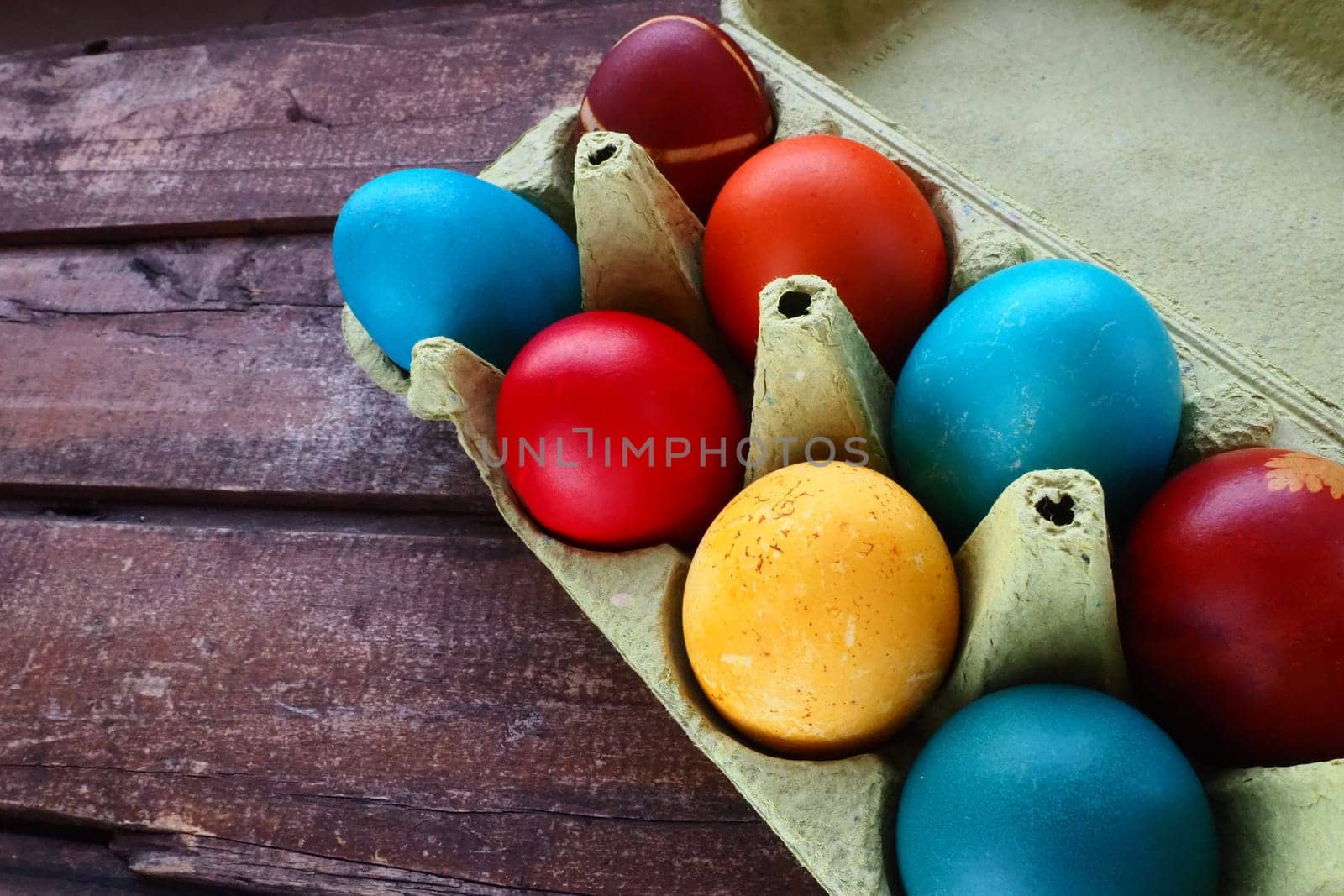 Multi-colored eggs painted with gouache and onion peel in a cardboard box on a wooden table background. Ten boiled eggs. Postcard poster for Easter. Easter holiday. Red, blue, yellow, brown eggs.