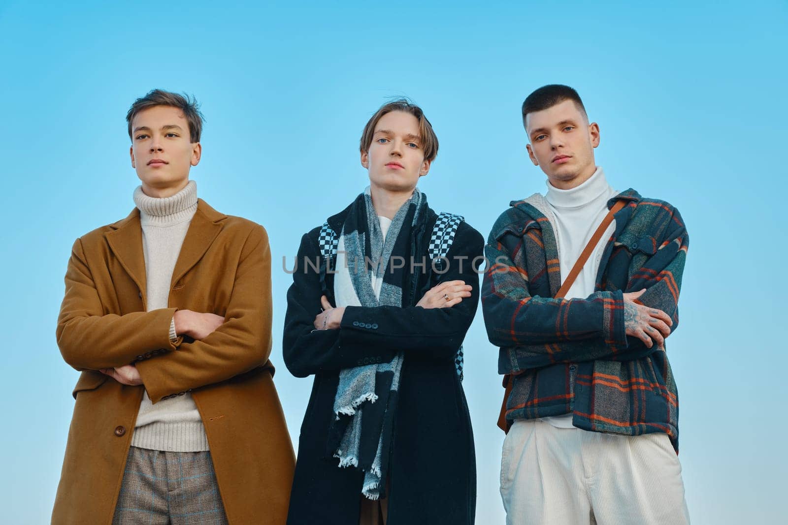 Three young men in autumn clothes are standing with crossed hands on the street