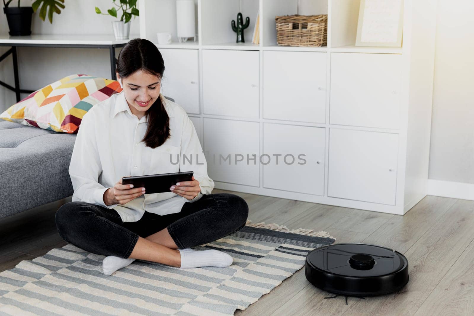 Young Caucasian woman using tablet, chatting, playing while robot vacuum cleaner is cleaning floor in the living room. Pretty girl is configuring device using mobile application over wifi network