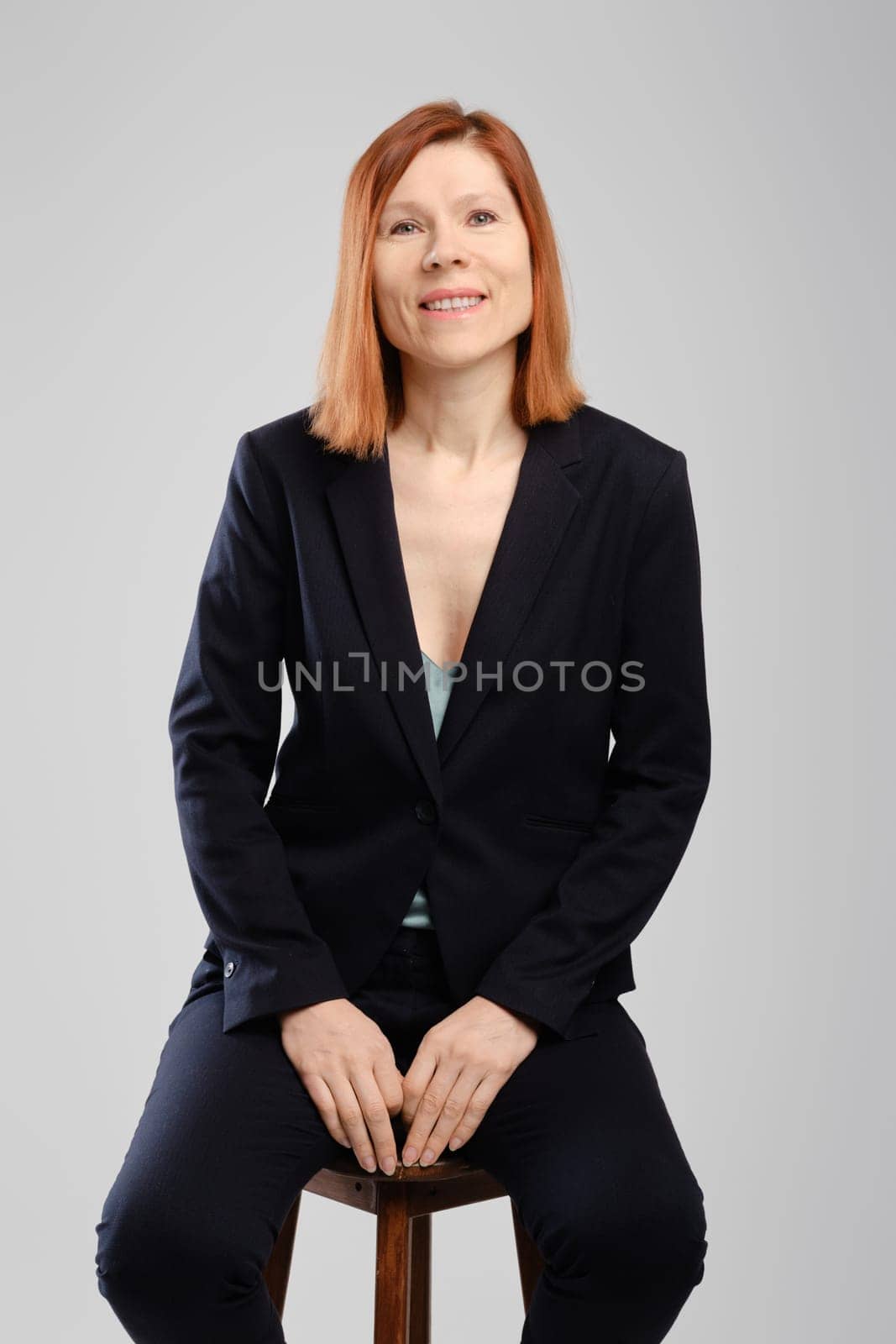 Stylish middle aged woman in pantsuit sitting in studio over grey background