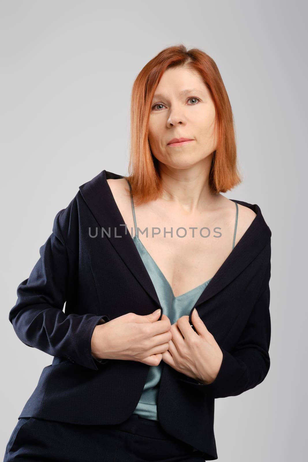 Stylish middle aged woman in pantsuit posing in studio holding arms on chest over grey background