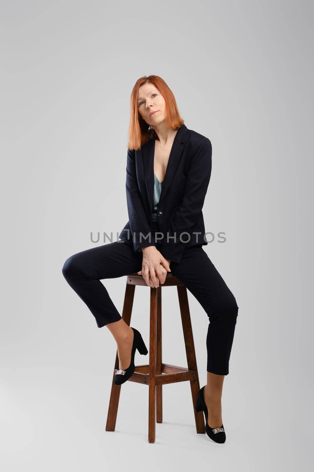 Stylish middle aged woman in pantsuit posing in studio sitting on chair with legs spread over grey background
