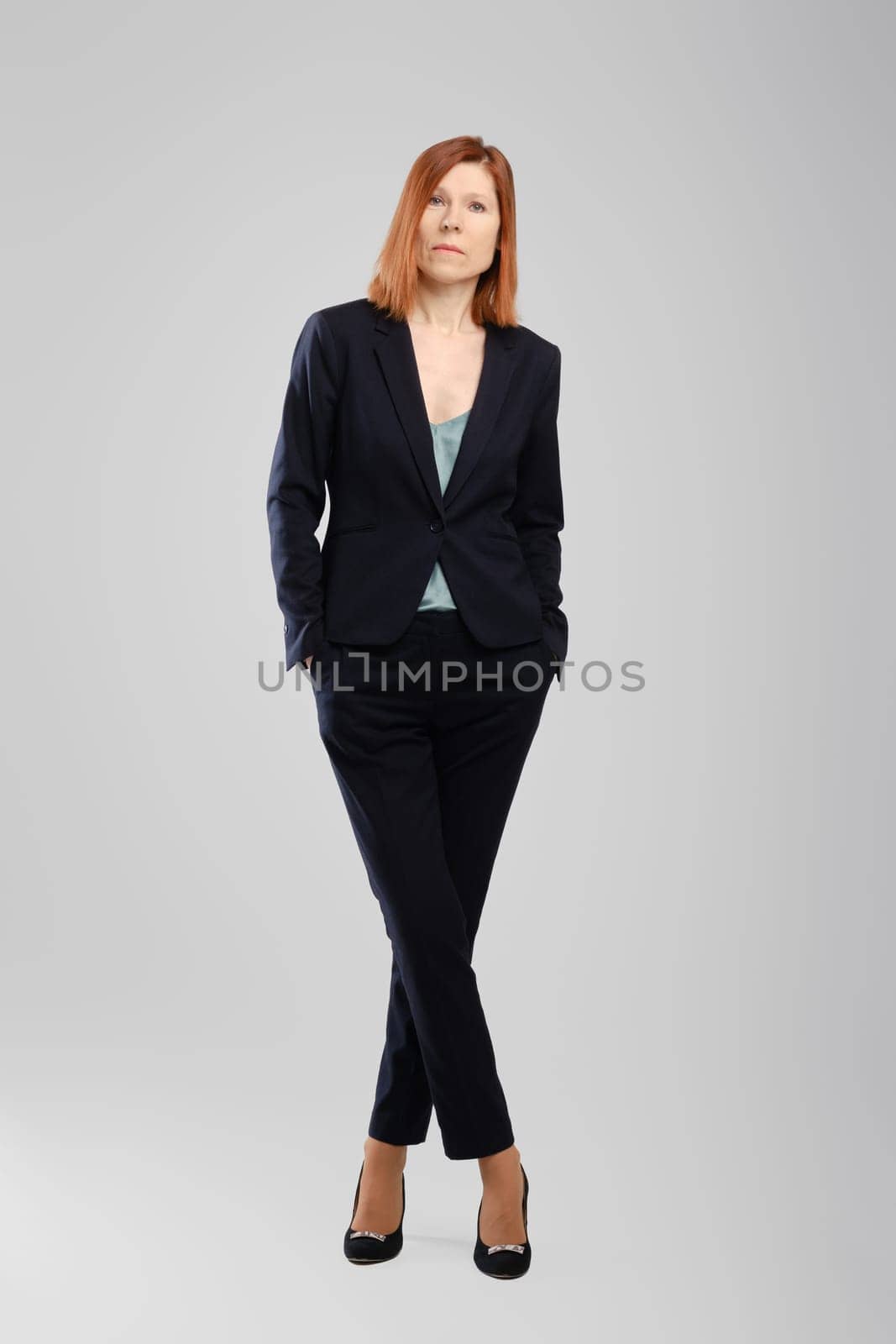 Stylish middle aged woman in pantsuit standing in studio over grey background
