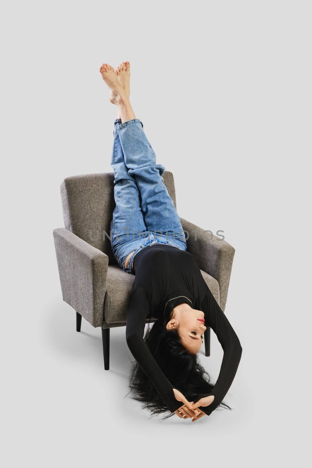 Young barefoot woman lays upside down in armchair in studio in black bodysuit with long sleeves and oversized jean over grey background