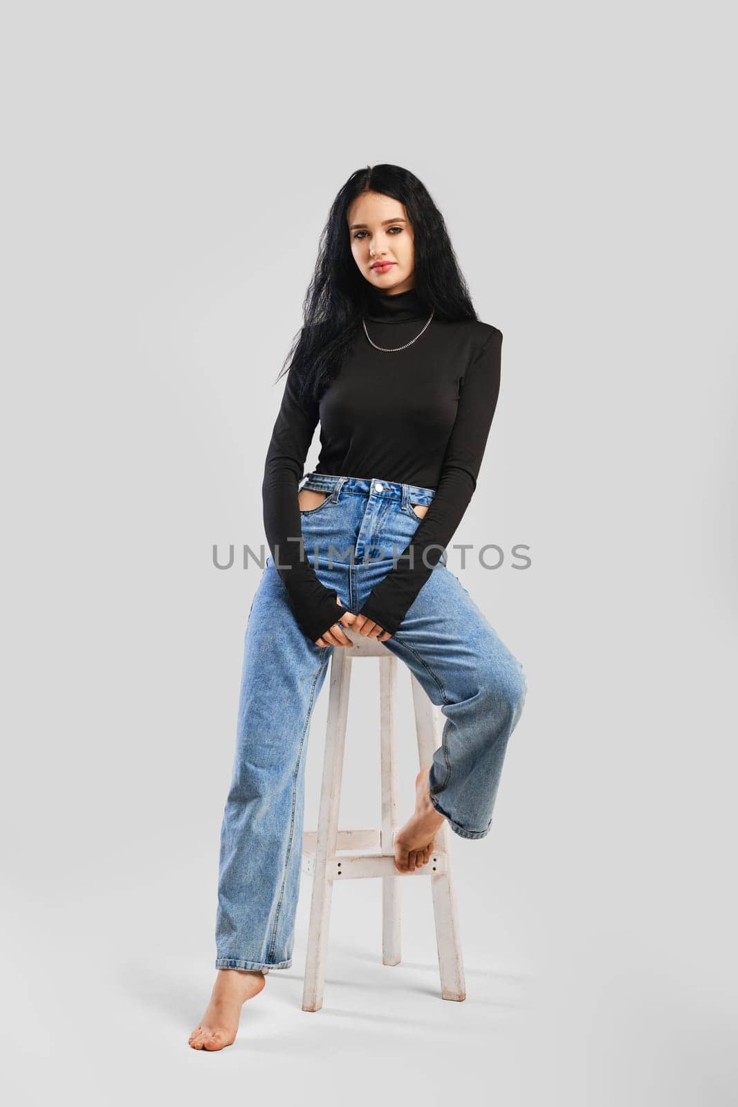 Young barefoot woman sits on chair in studio in black bodysuit with long sleeves and oversized jean over grey background