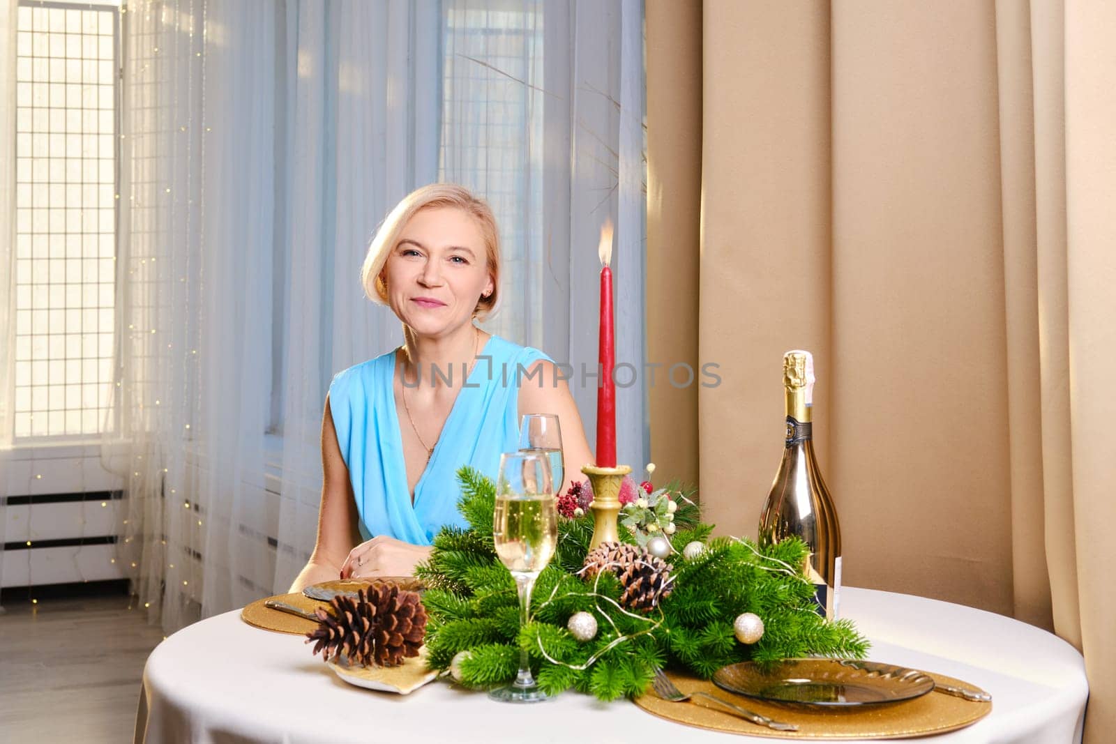 Charming middle age woman in long blue dress sits behind the festive christmas table at home