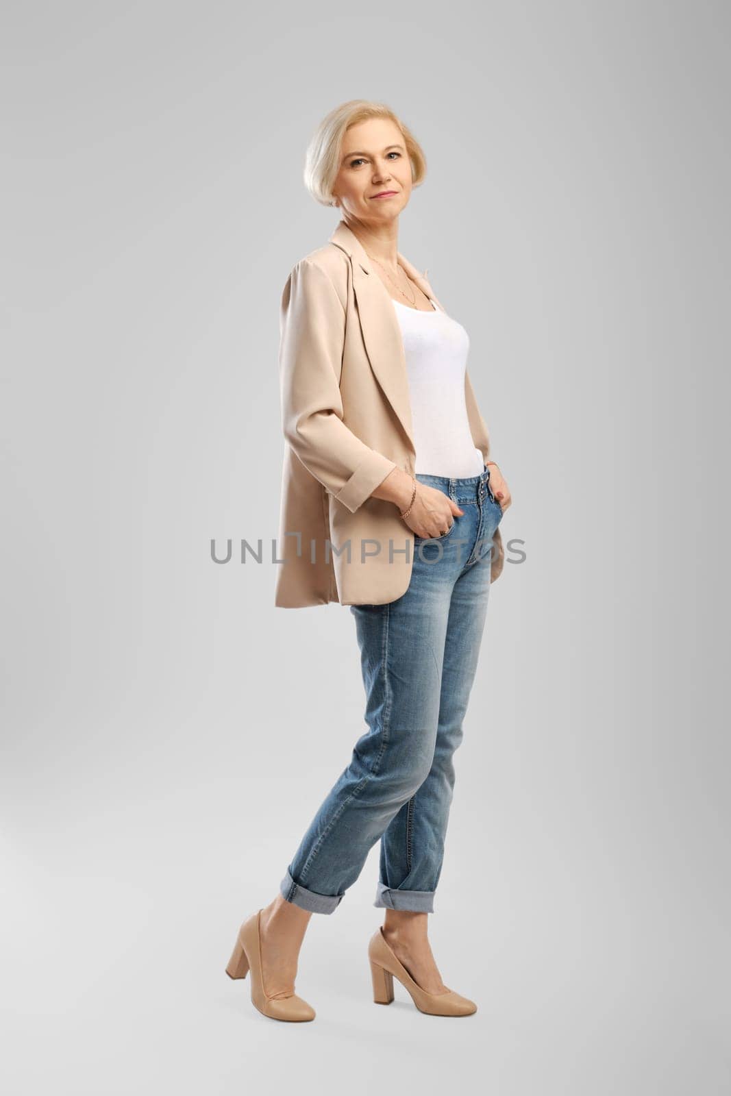 Full length portrait of senior woman in white tank top, beige jacket and boyfriend jeans over grey background