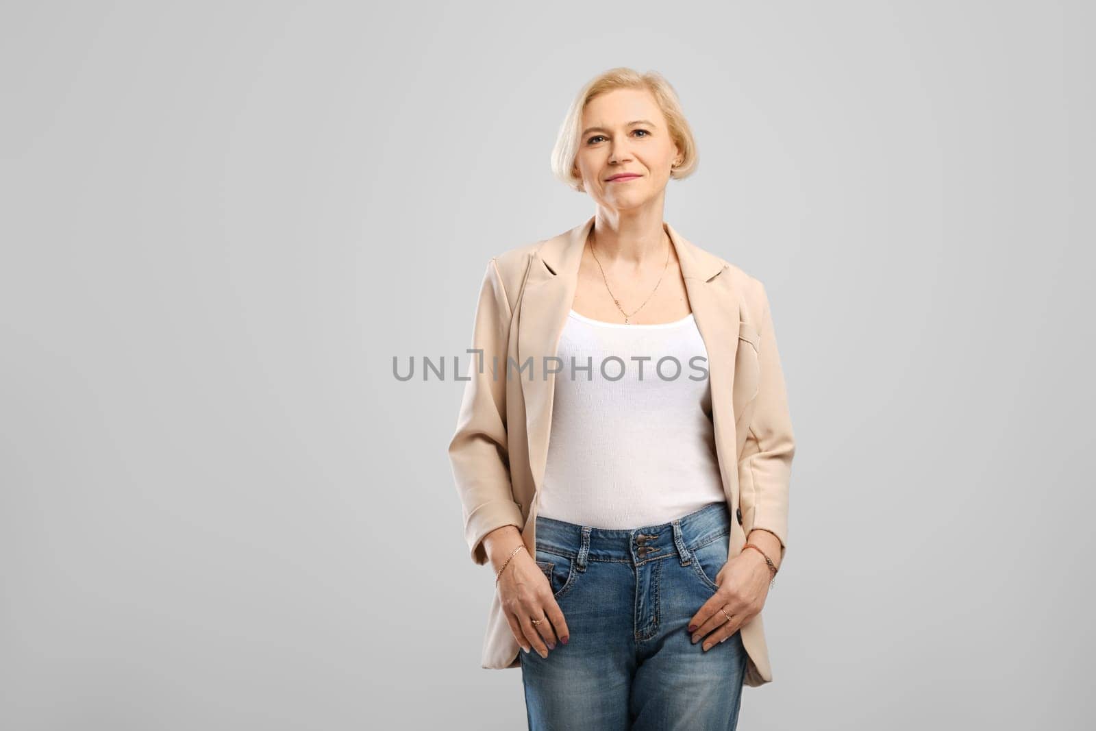 Confident middle age woman standing with arms in the pockets looking at camera at gray background