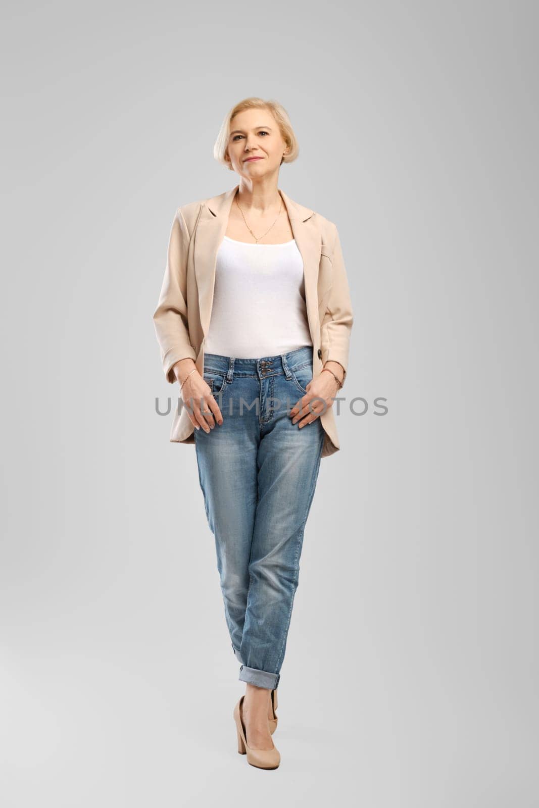Senior woman in white tank top, beige jacket and boyfriend jeans walking in studio over grey background