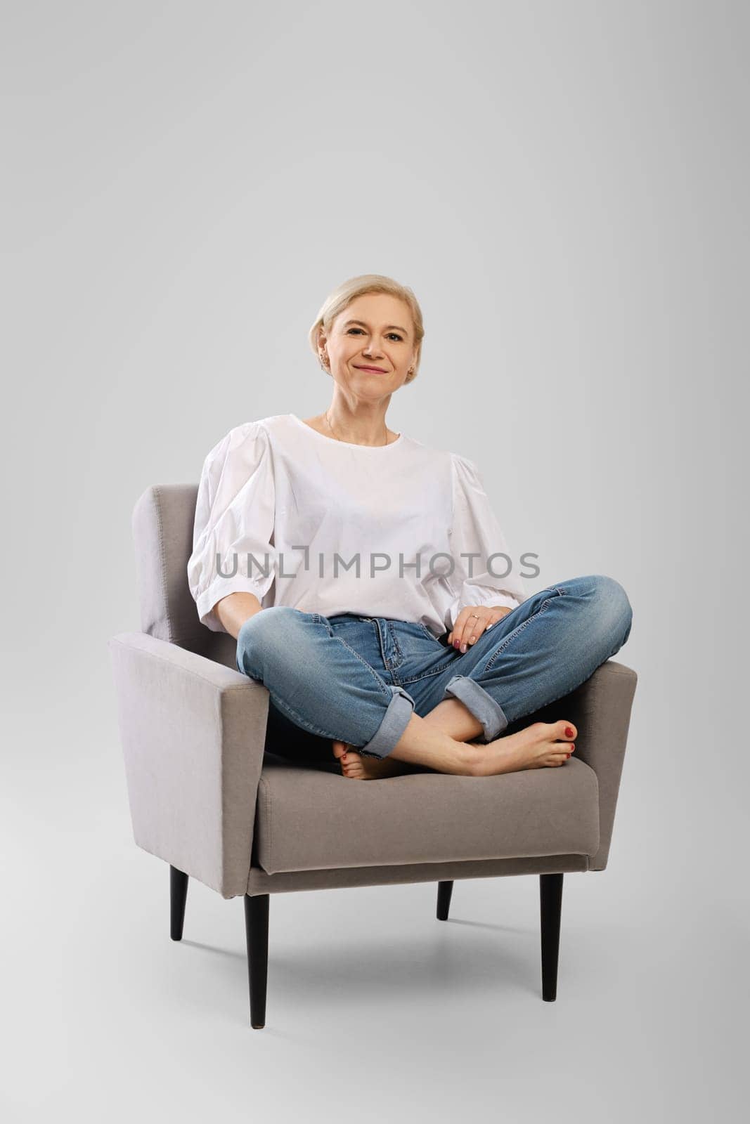 Barefoot senior woman sitting in an armchair crossing legs in studio