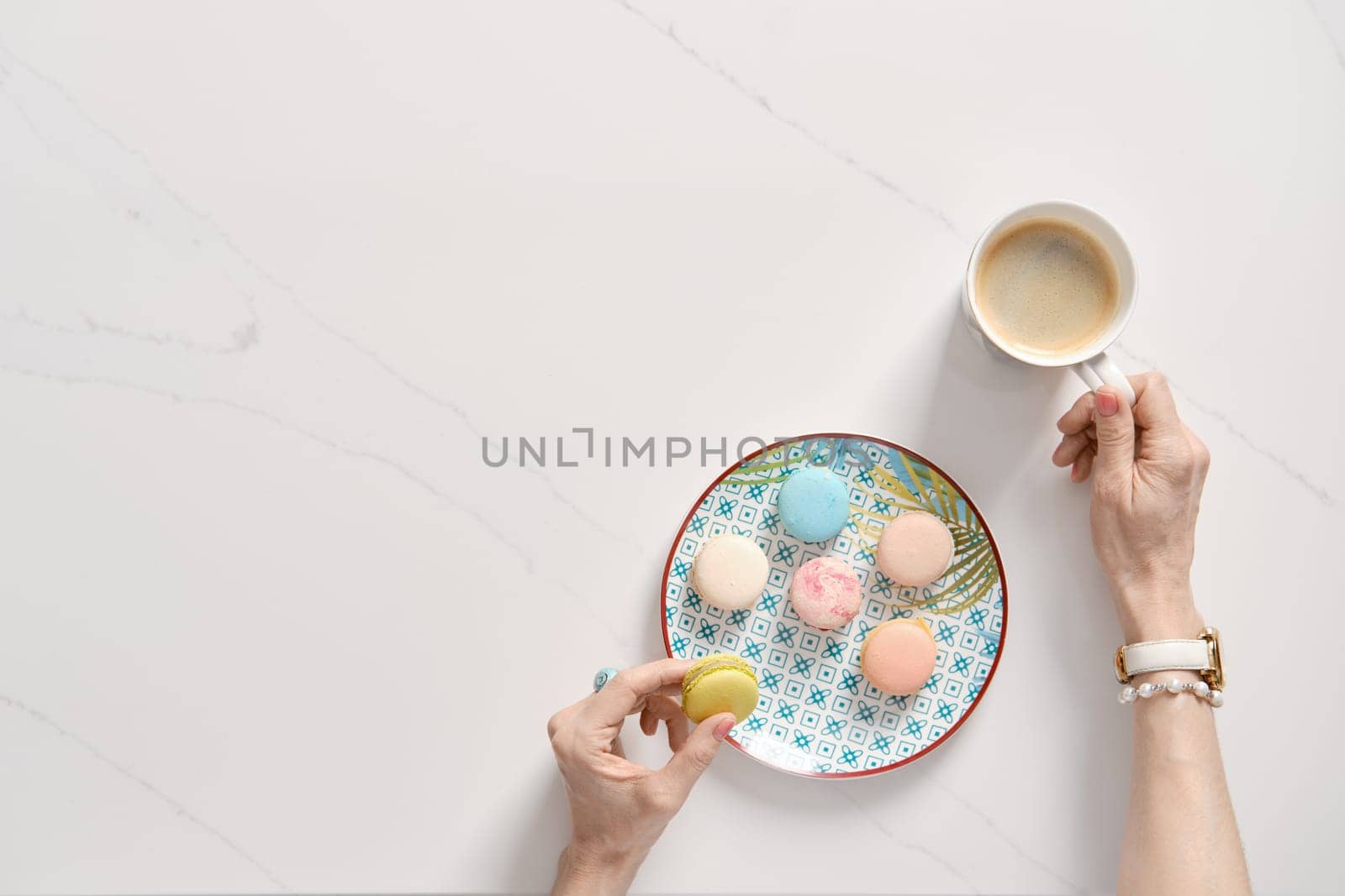 Top view of colorful cake macaroons on a plate and female hand with cup of coffee