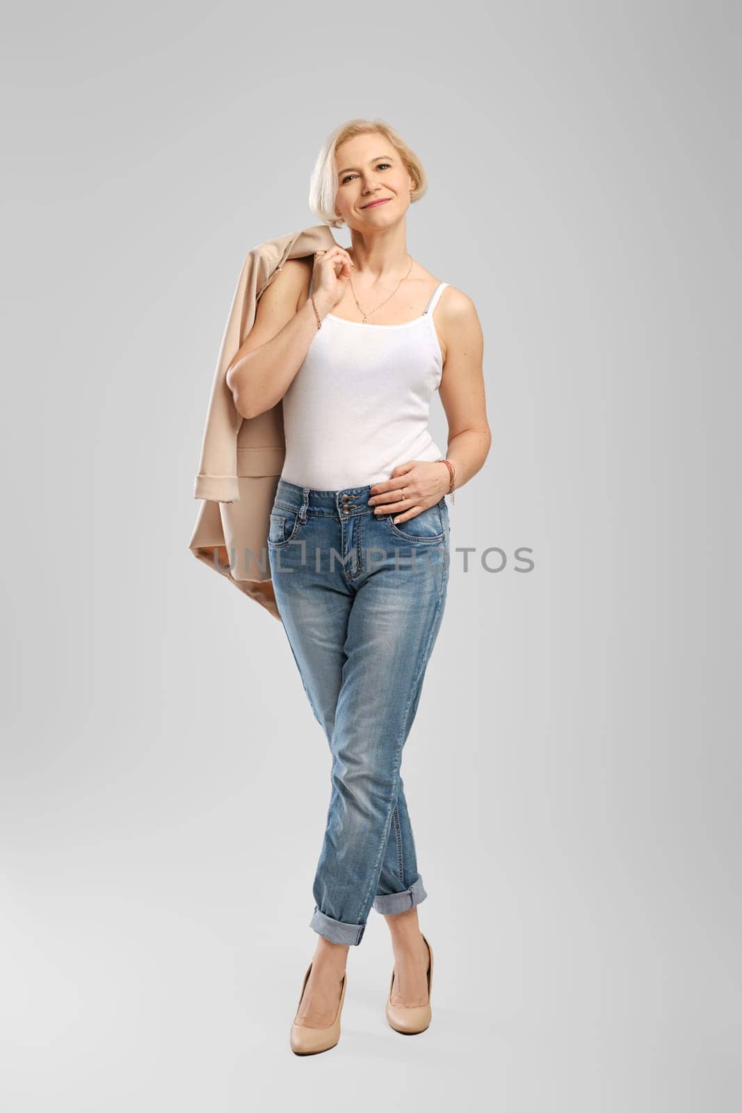 Full length portrait of senior woman in white tank top holding beige jacket over the shoulder in studio