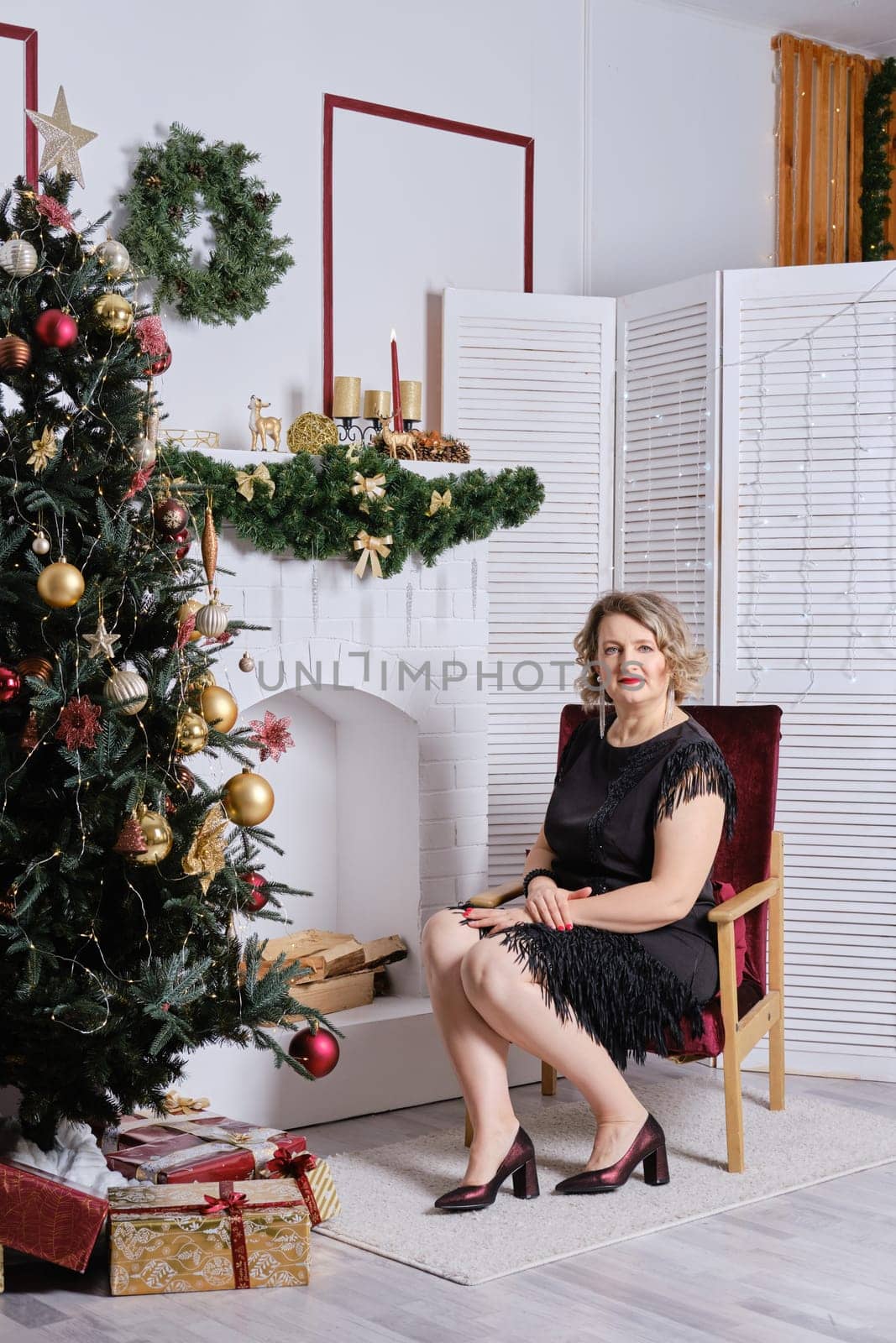 Middle age woman sitting in cosy armchair near christmas tree and fireplace