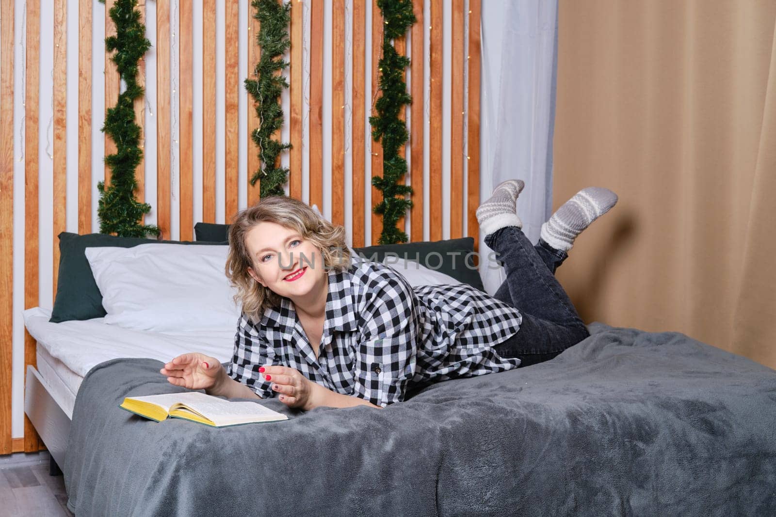 Cheerful middle age woman in casual wear lays on bed and reading book