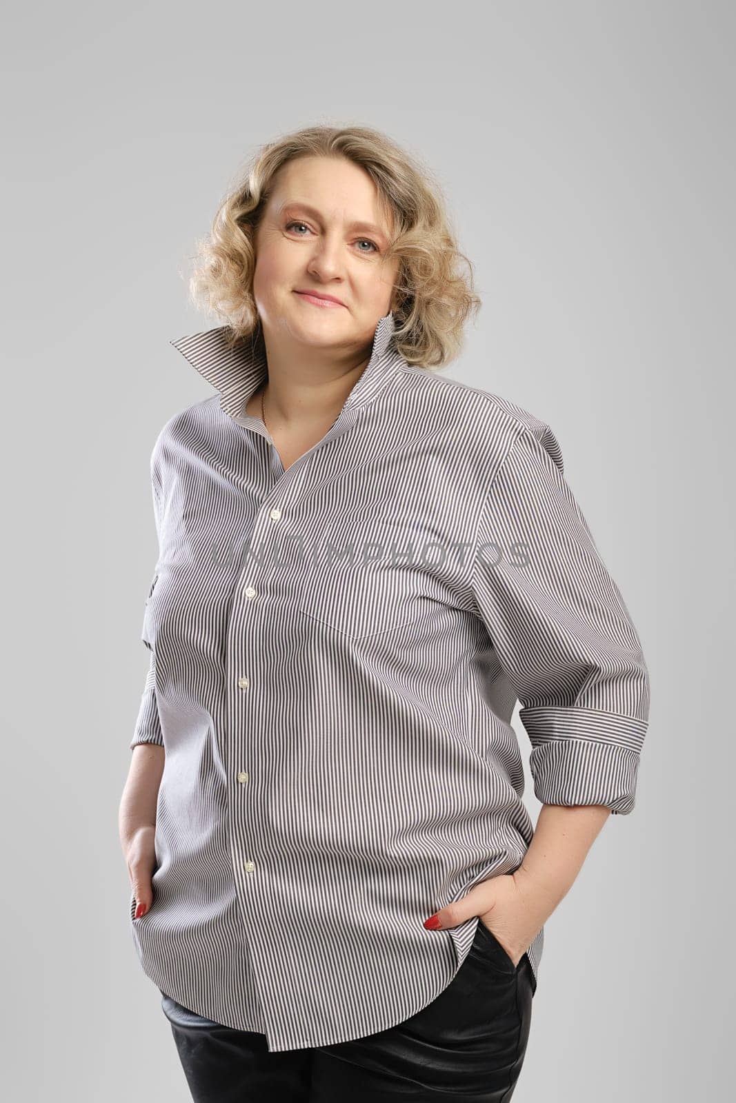 Casual portrait of senior woman in grey shirt and trousers with hands in pockets over grey studio background