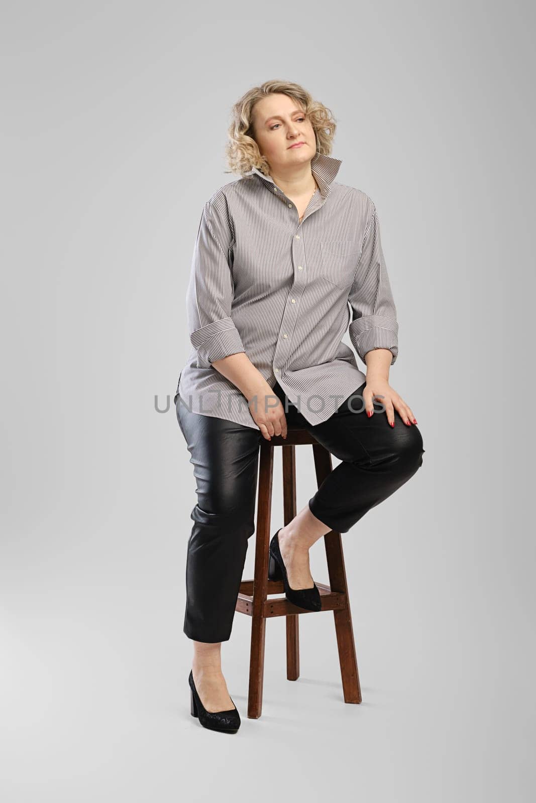 Senior woman in grey shirt and faux leather trousers sits on tall wooden chair over grey studio background