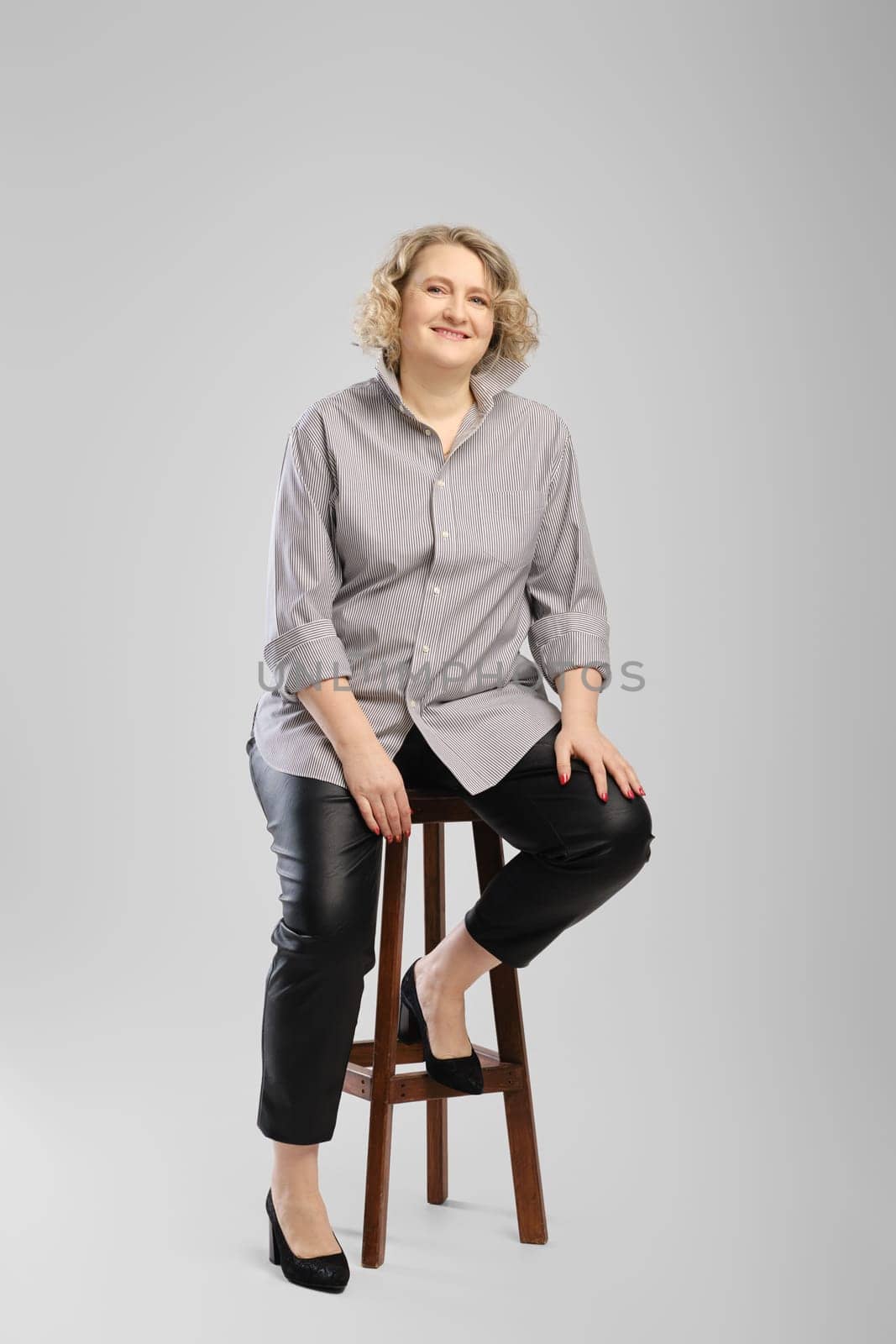 Senior woman in grey shirt and faux leather trousers sits on tall wooden chair over grey studio background