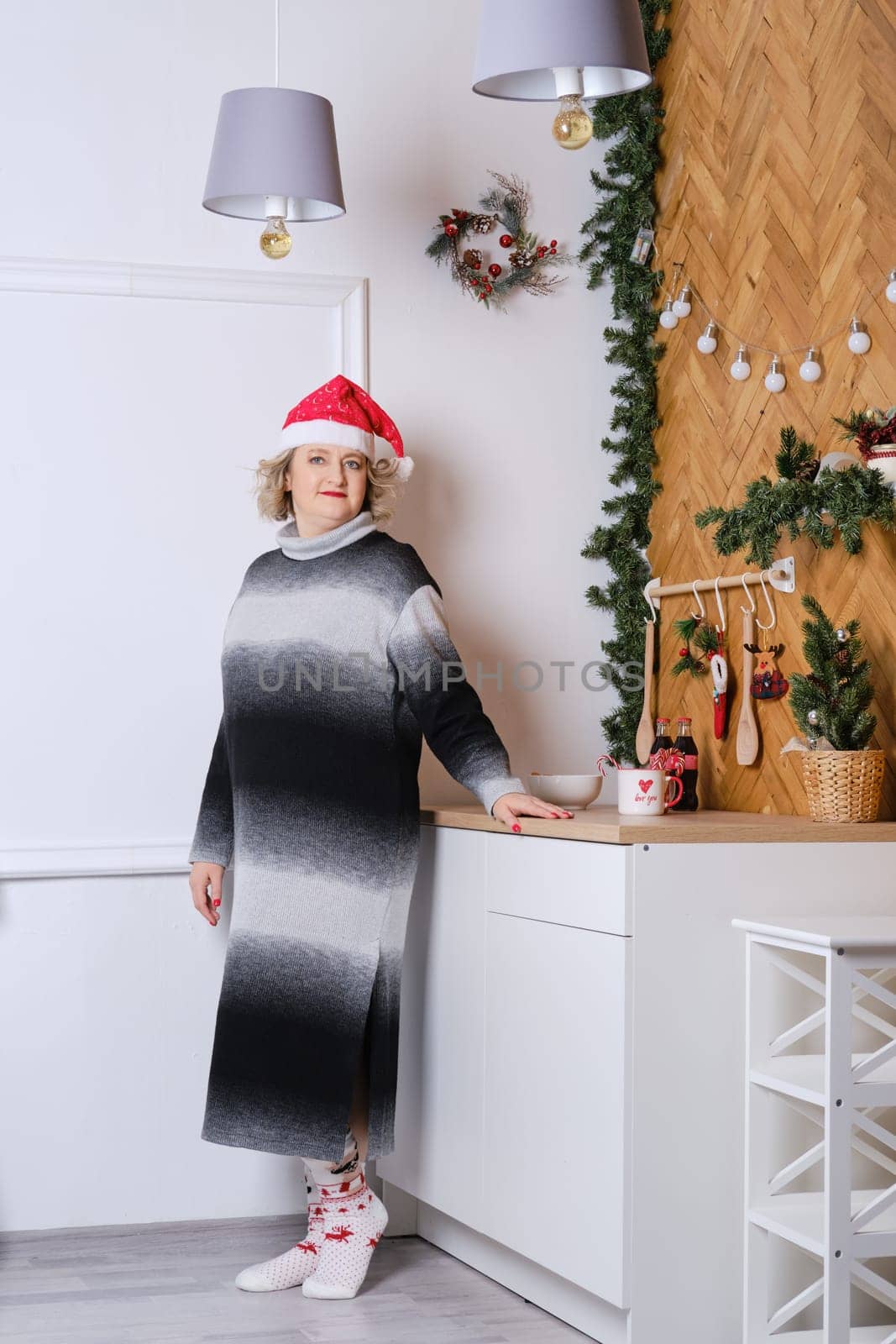 Full length portrait of middle age woman in christmas cap standing in small kitchen and holds a cup with hot beverage in hand