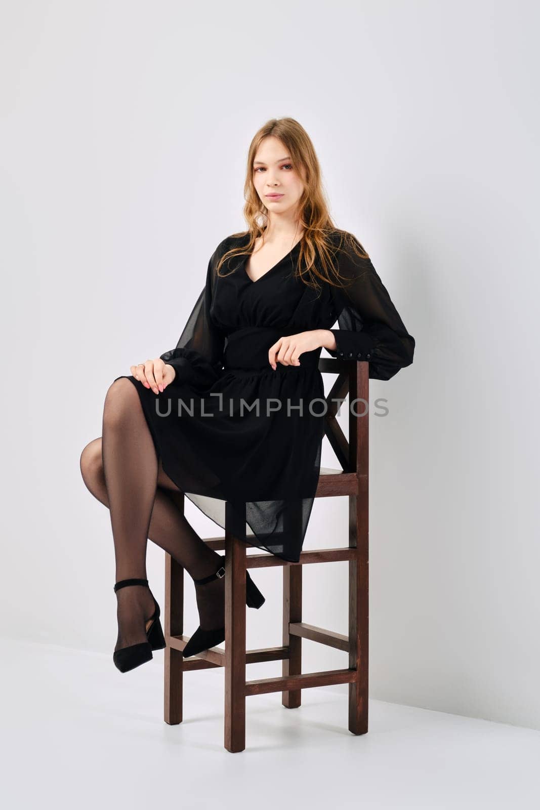 Full length portrait of cute young woman in black tulle dress sitting in profile on tall chair in white studio
