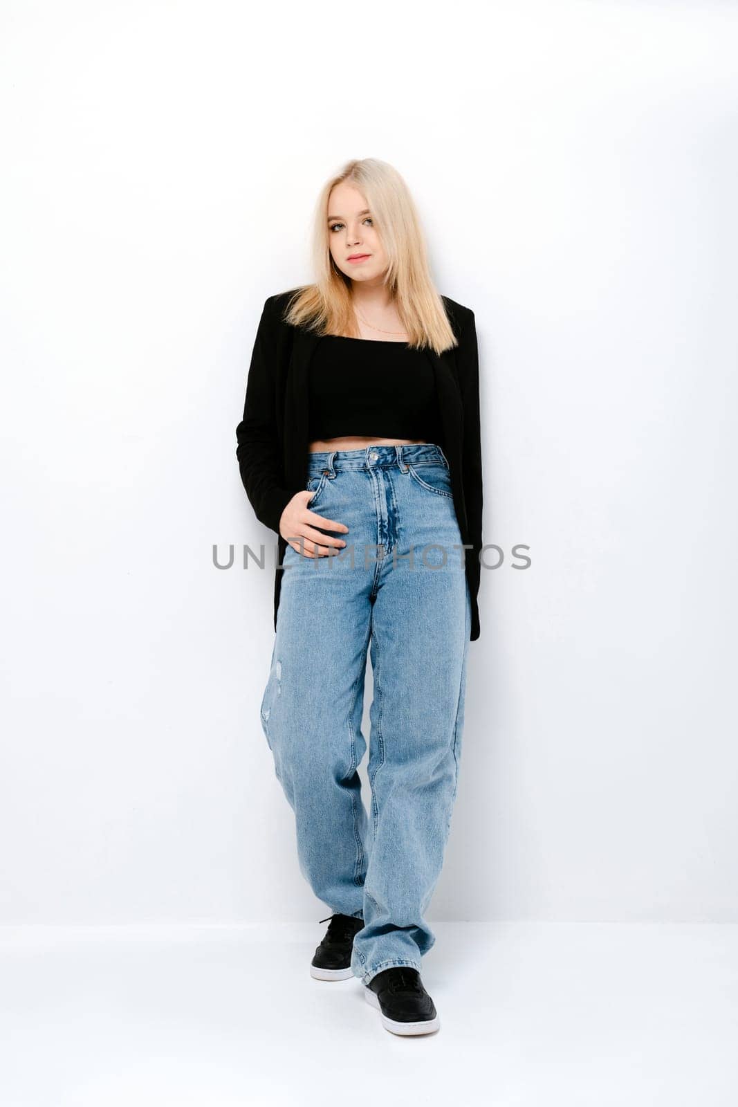 Blonde girl in tank black top, jacket and jeans posing over white wall