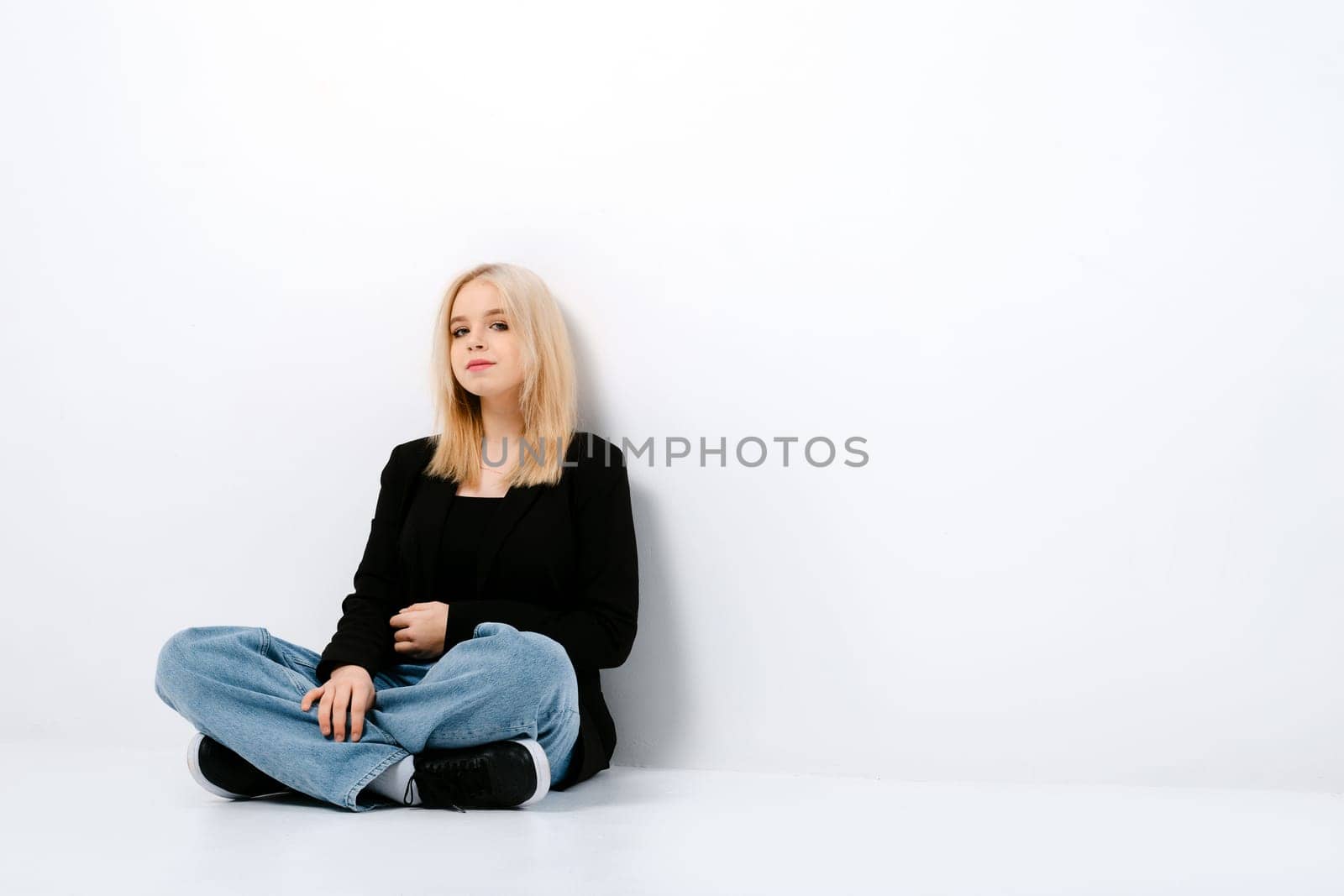 Pensive girl in tank top, jacket and jeans posing sitting on the floor with crossed legs in studio