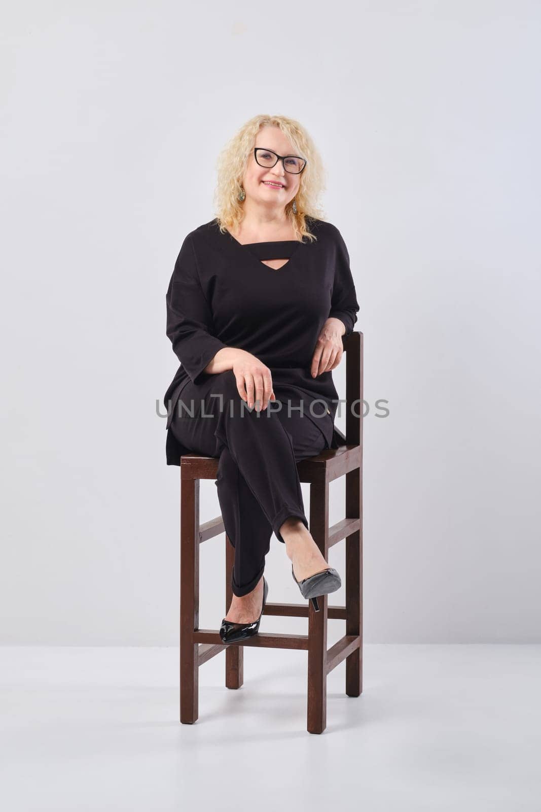Middle age woman in black outfit and glasses on chair, studio shot