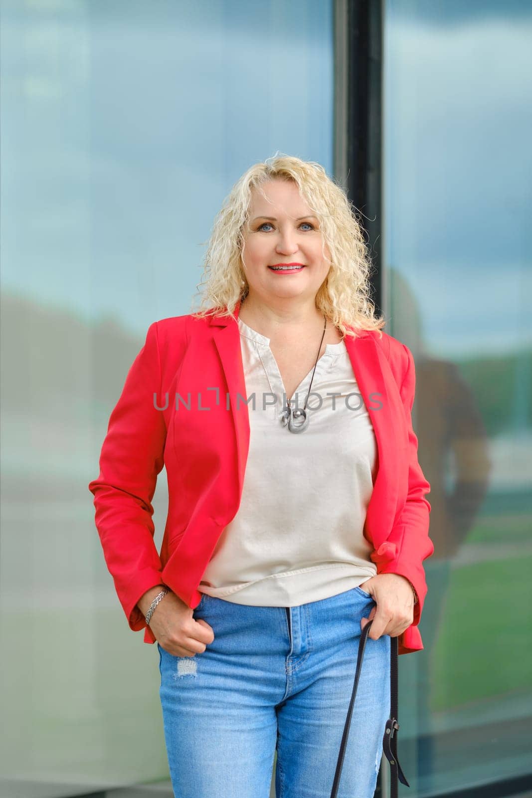 woman in red jacket, blouse and denim jeans standing near the reinforced glass wall of the building