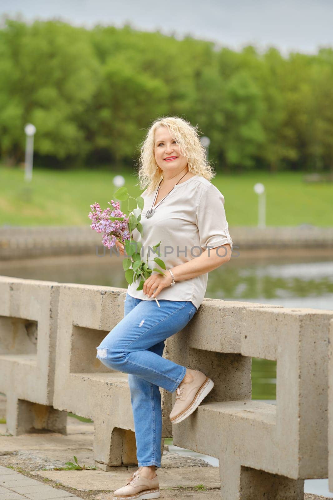 Smiling senior woman walks along the embankment with a branch of lilac in her hands