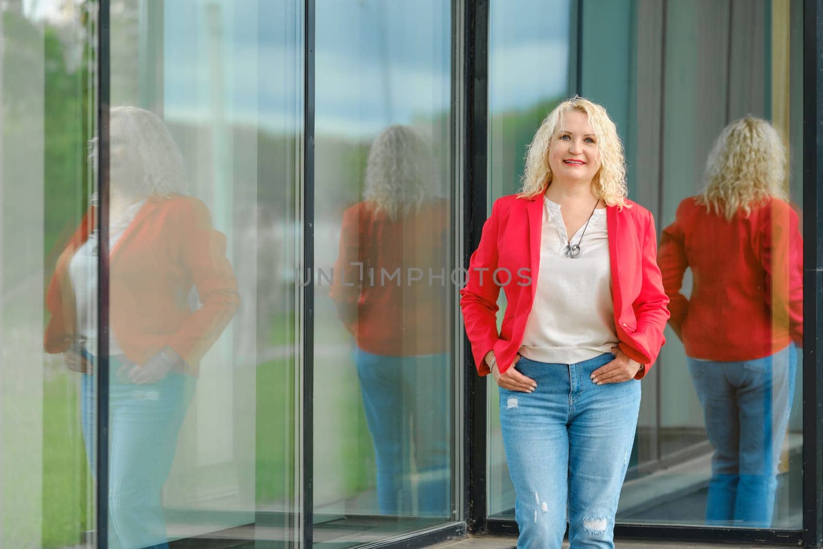 Cute senior woman in red jacket, blouse and denim jeans standing near the tempered glass wall of the building