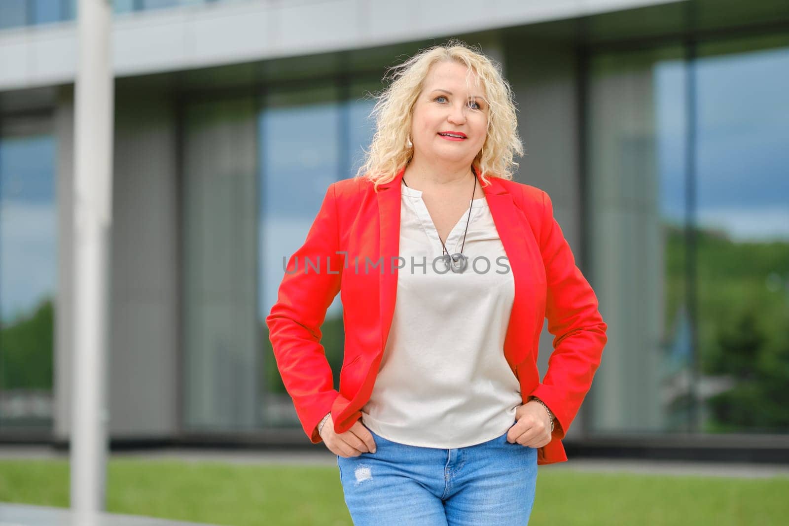 Confident senior woman in red jacket, blouse and denim jeans standing near the modern building