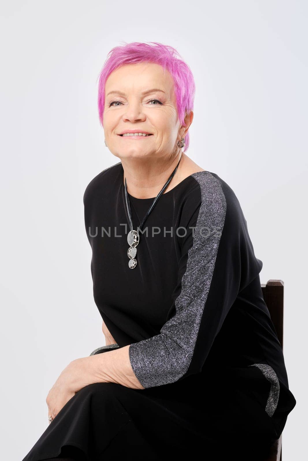Cheerful old woman with short pink hair sitting leaning on her knee over white background